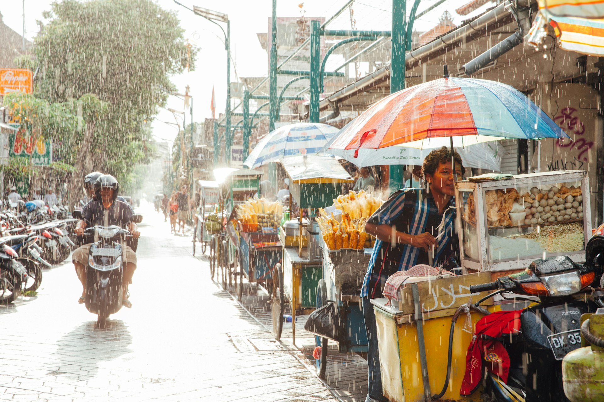 A motorcycle with a passenger drives down a street lined with food stalls in the rain as the sun breaks through the clouds