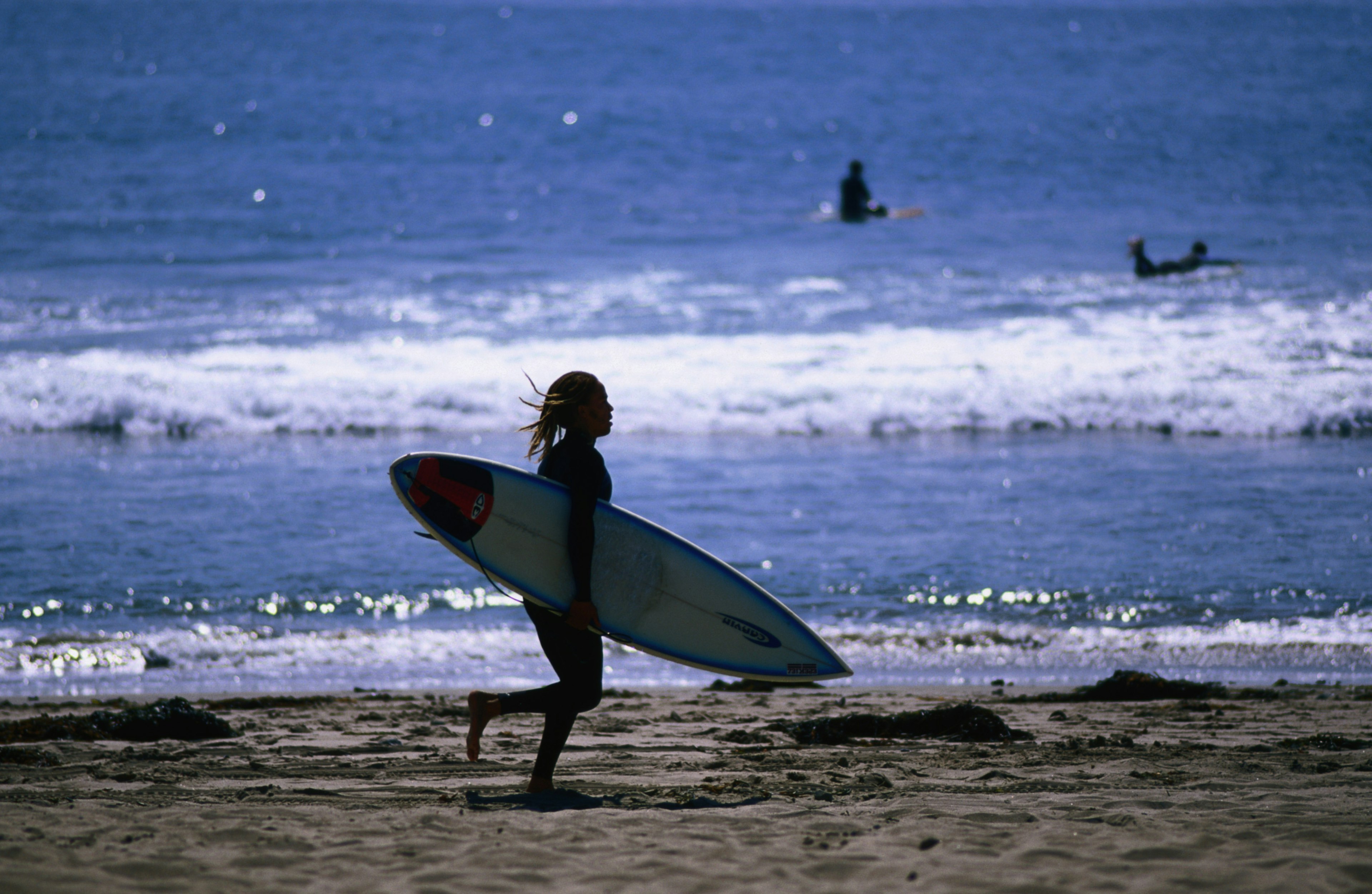 A surfer with long hair runs along the beach holding onto a surf board