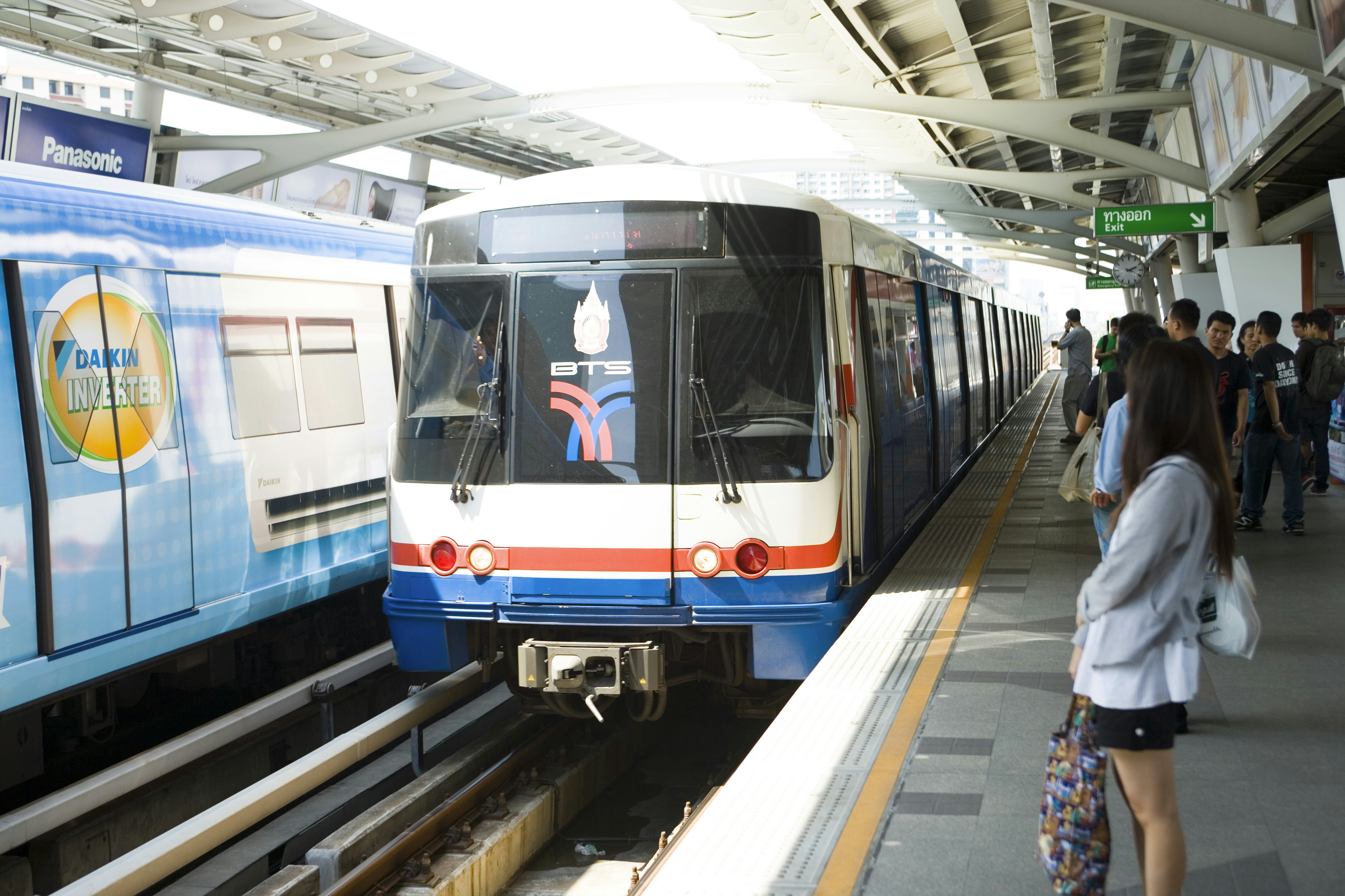 The blue, white and red BTS Skytrain pulls into Ari station in Bangkok with passengers moving towards it to embark.