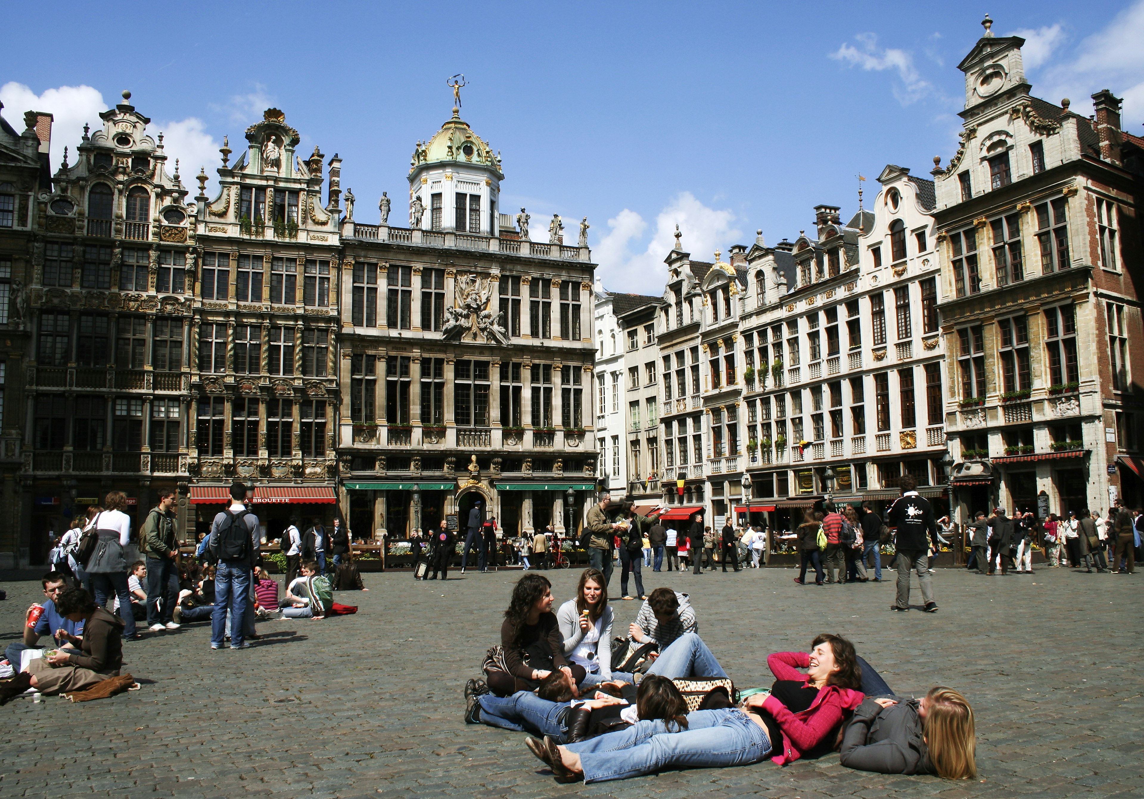 A group of people relax on the floor of the main square in Prague. Many other groups of people sit around them. The sun is shining and historic white buildings line the square.