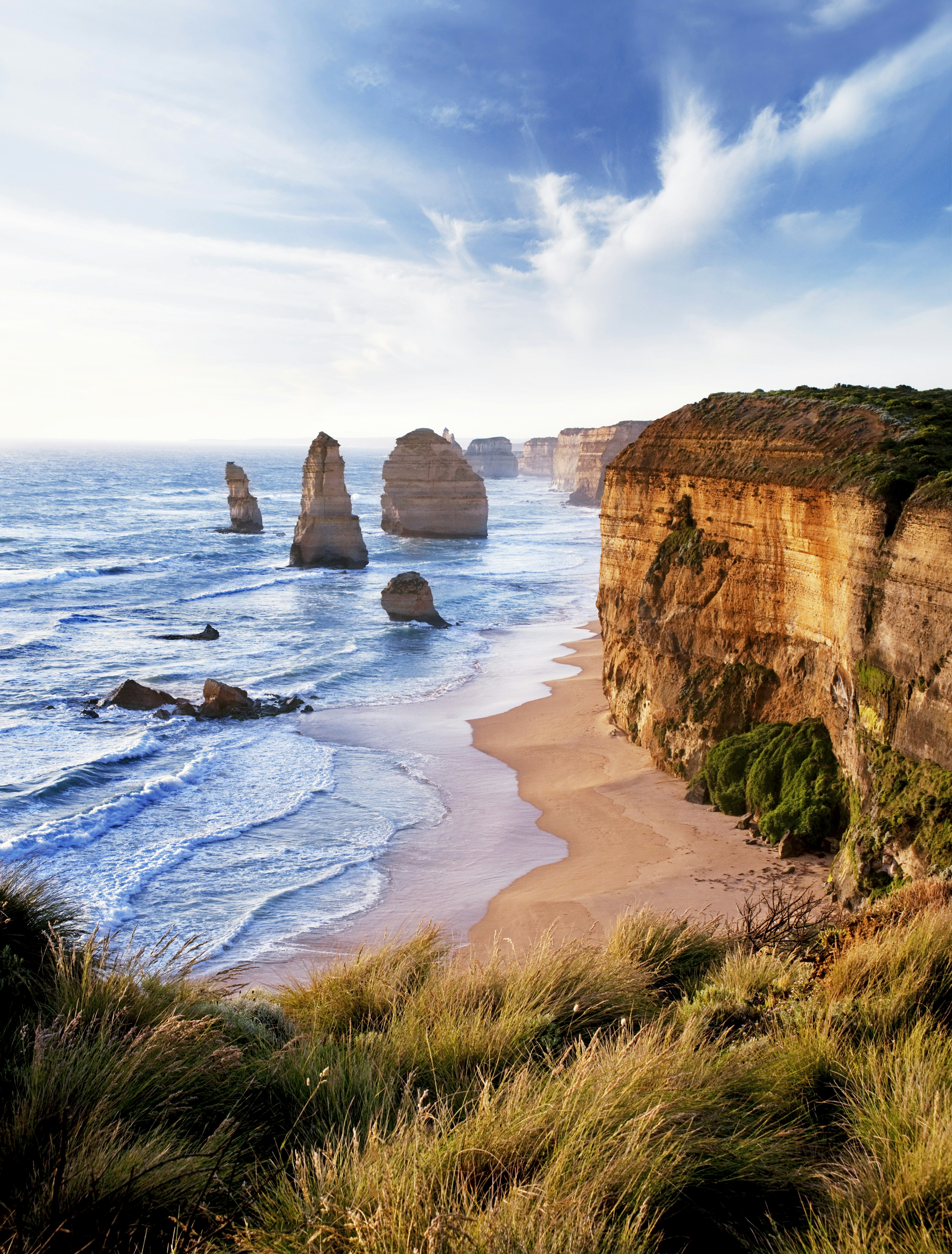 A sunset frames huge limestone rock formations sitting on the beach next to an undulating cliff line in Victoria, Australia.