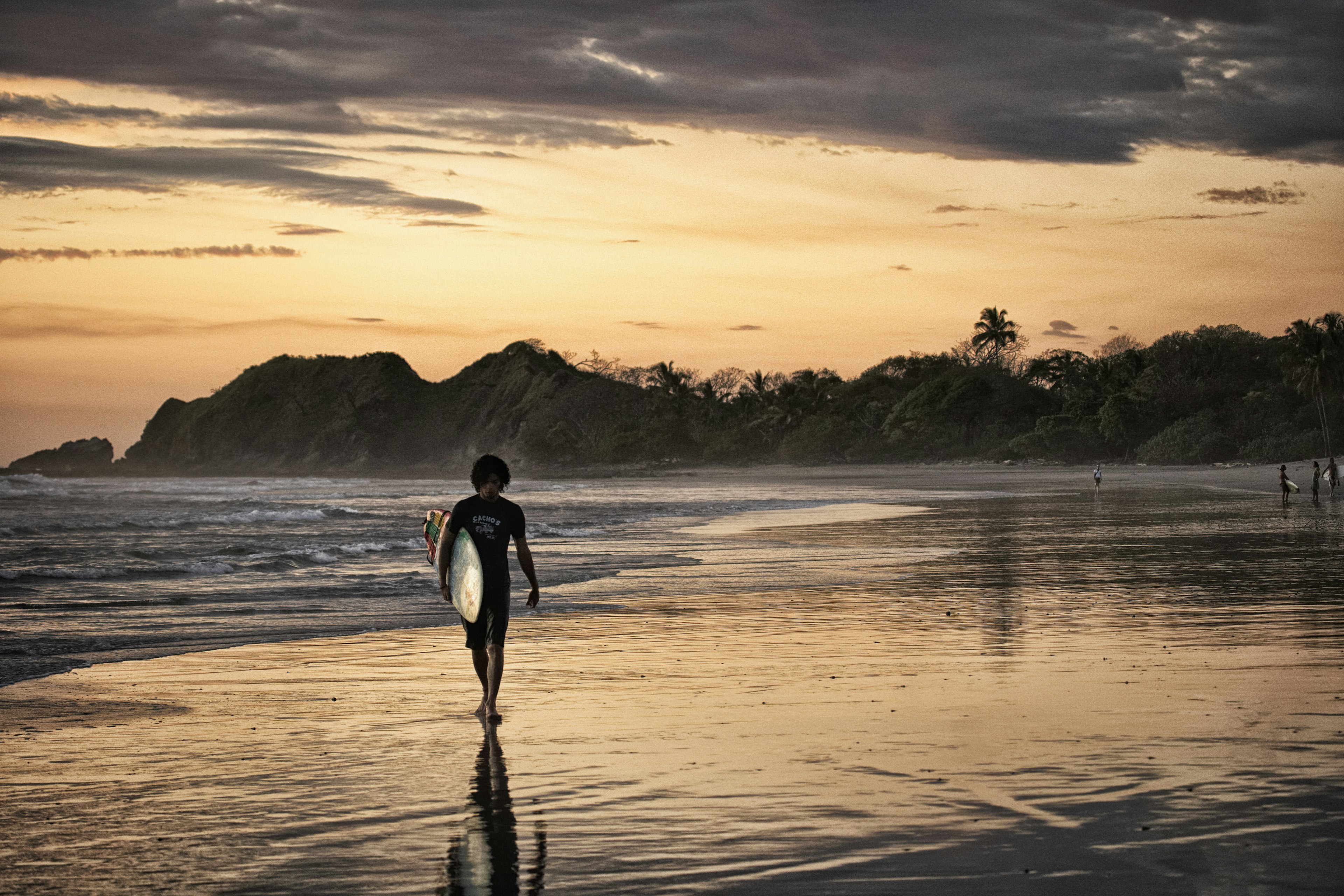 A surfer holding their board is silhouetted in the pink sunset light as they walk along a beach