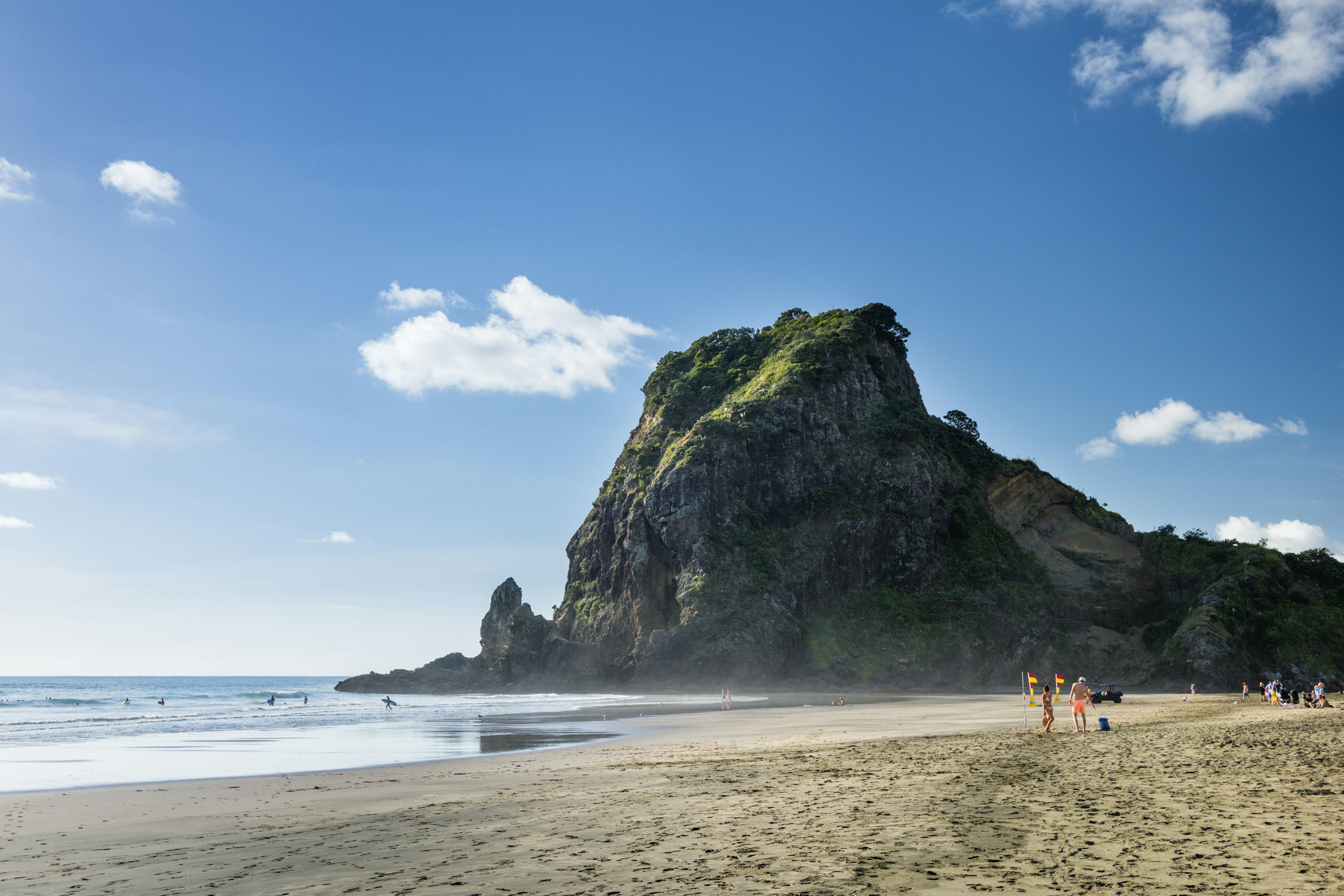 Swimmers and surfers enter the water between flags marking the safe area to swim, all under the shadow of a massive rocky outcrop