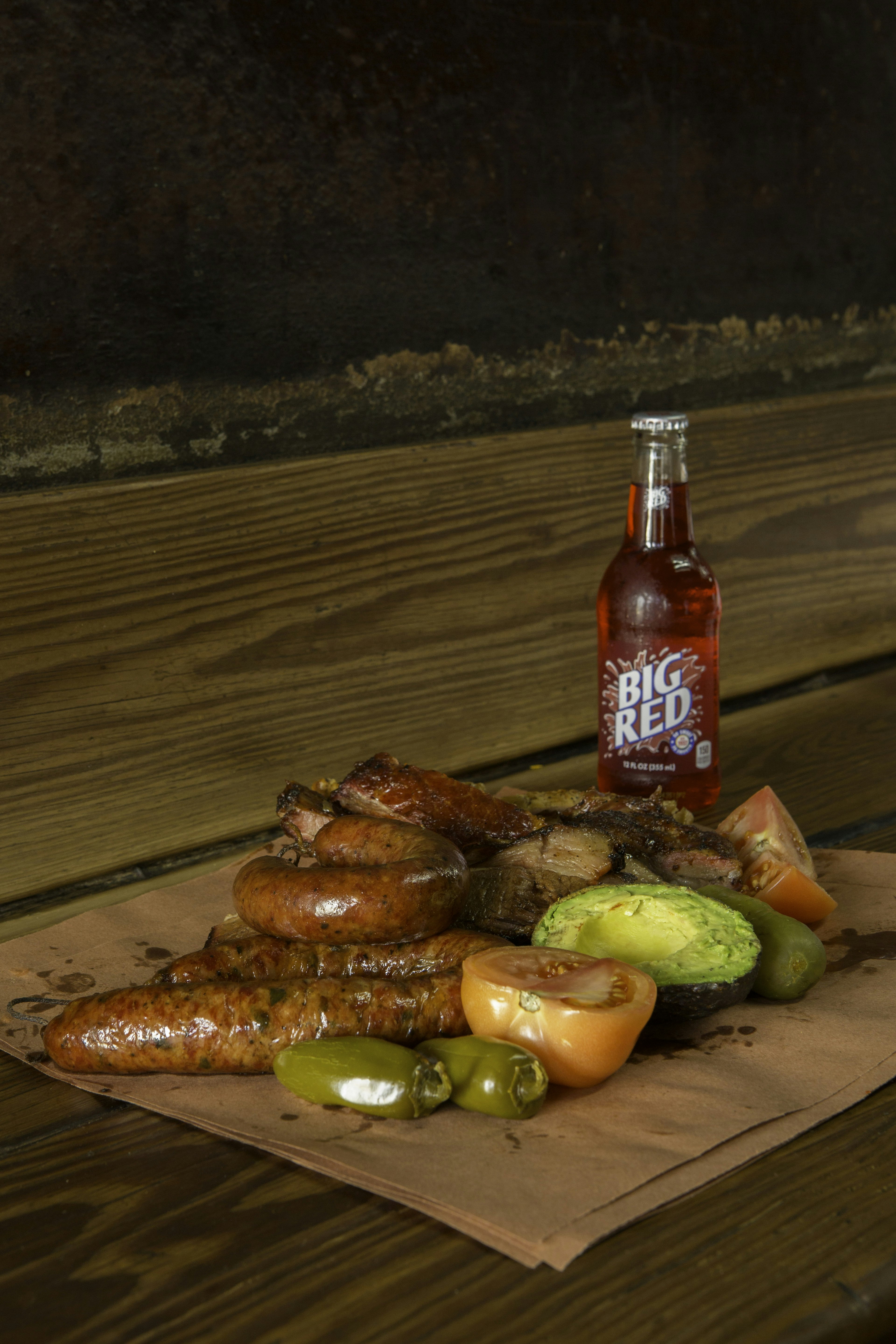 A piece of butcher paper piled with a collection of barbecue meats with tomatoes and peppers. There is a bottle of Big Red next to the food.