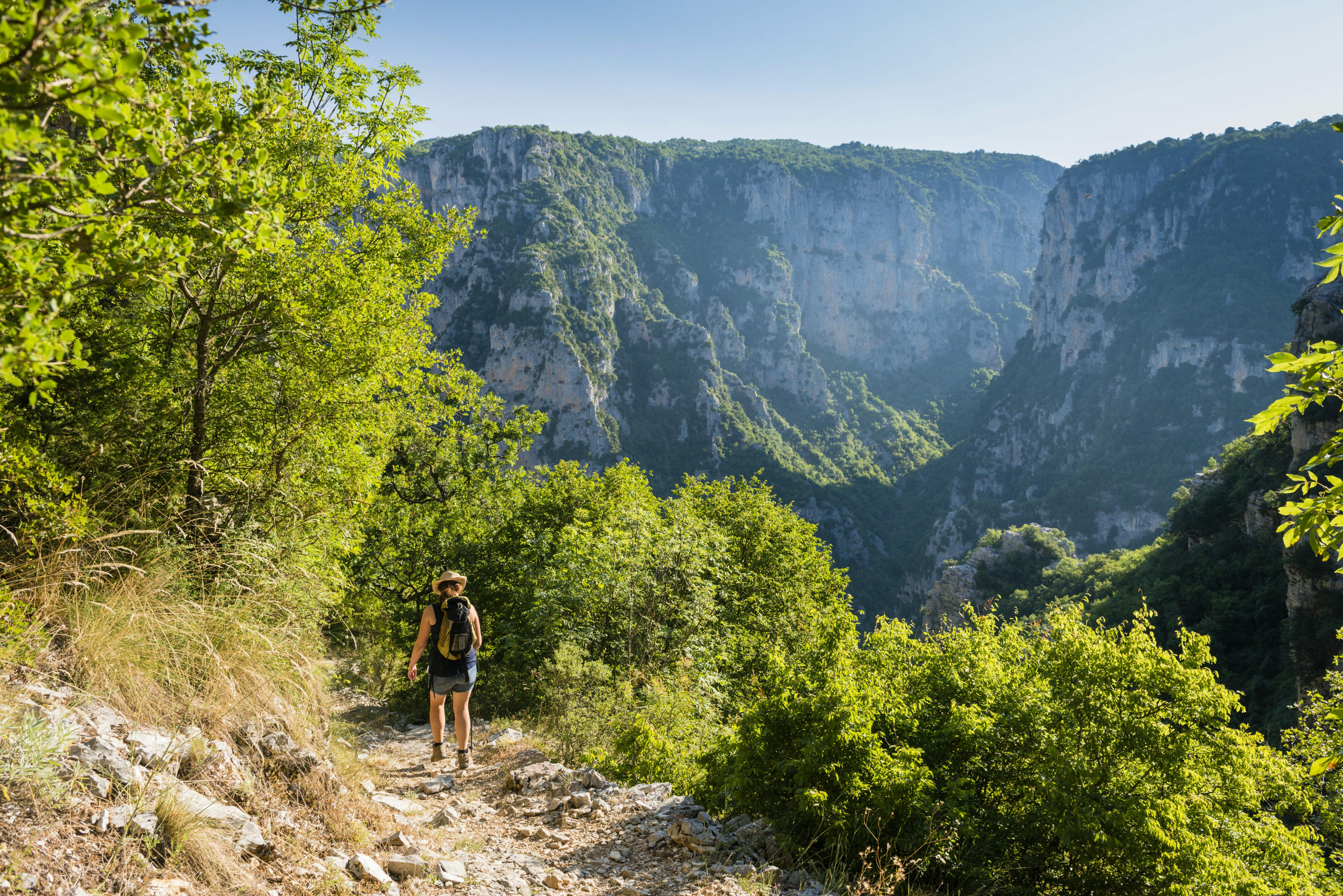 A hiker walks along a cliffside path into Vikos Gorge. The large valley gorge looms large in the background, with rocky vertical walls on either side.