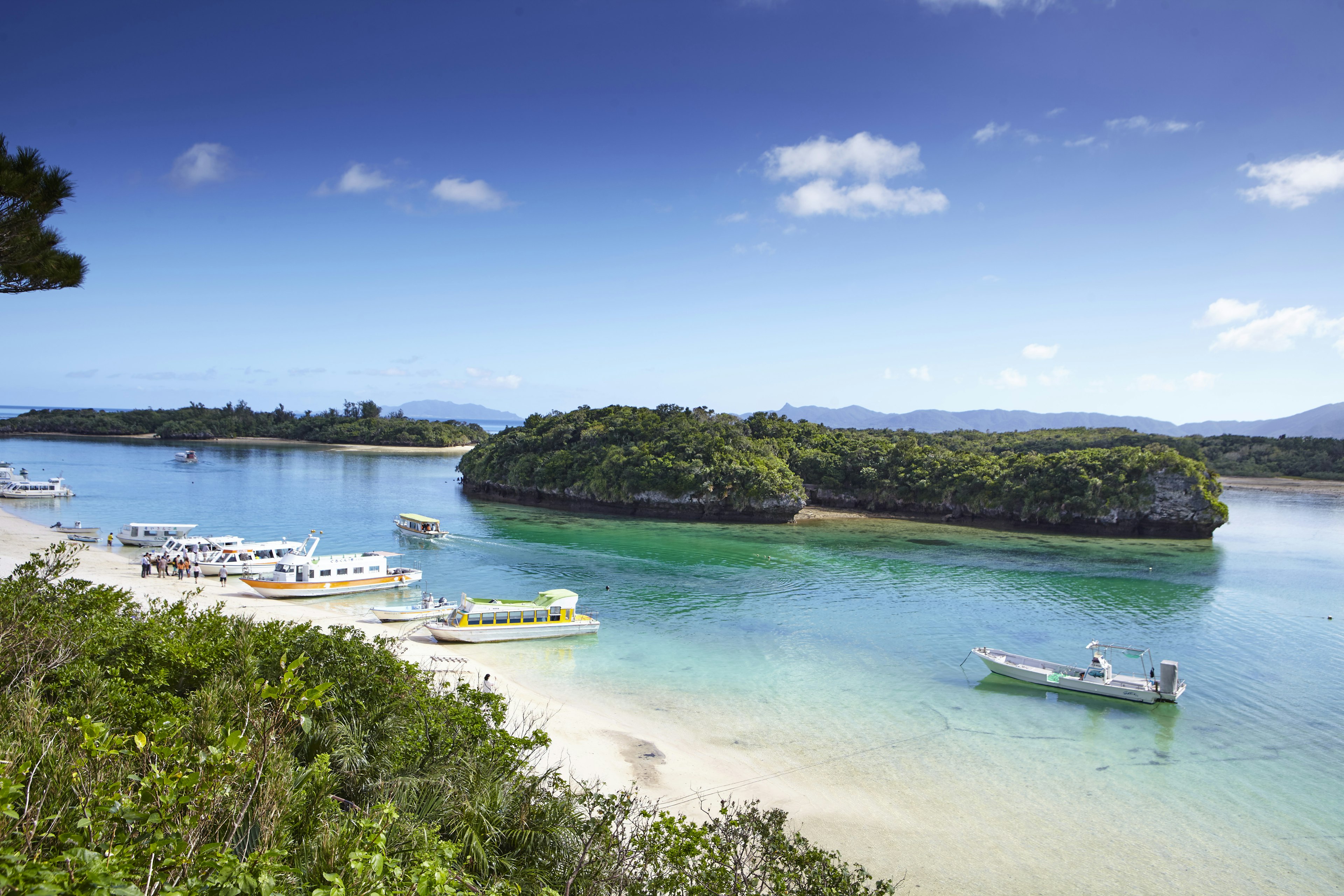 A white-sand beach lined with several small tourist boats; several rocky islets are in the bay