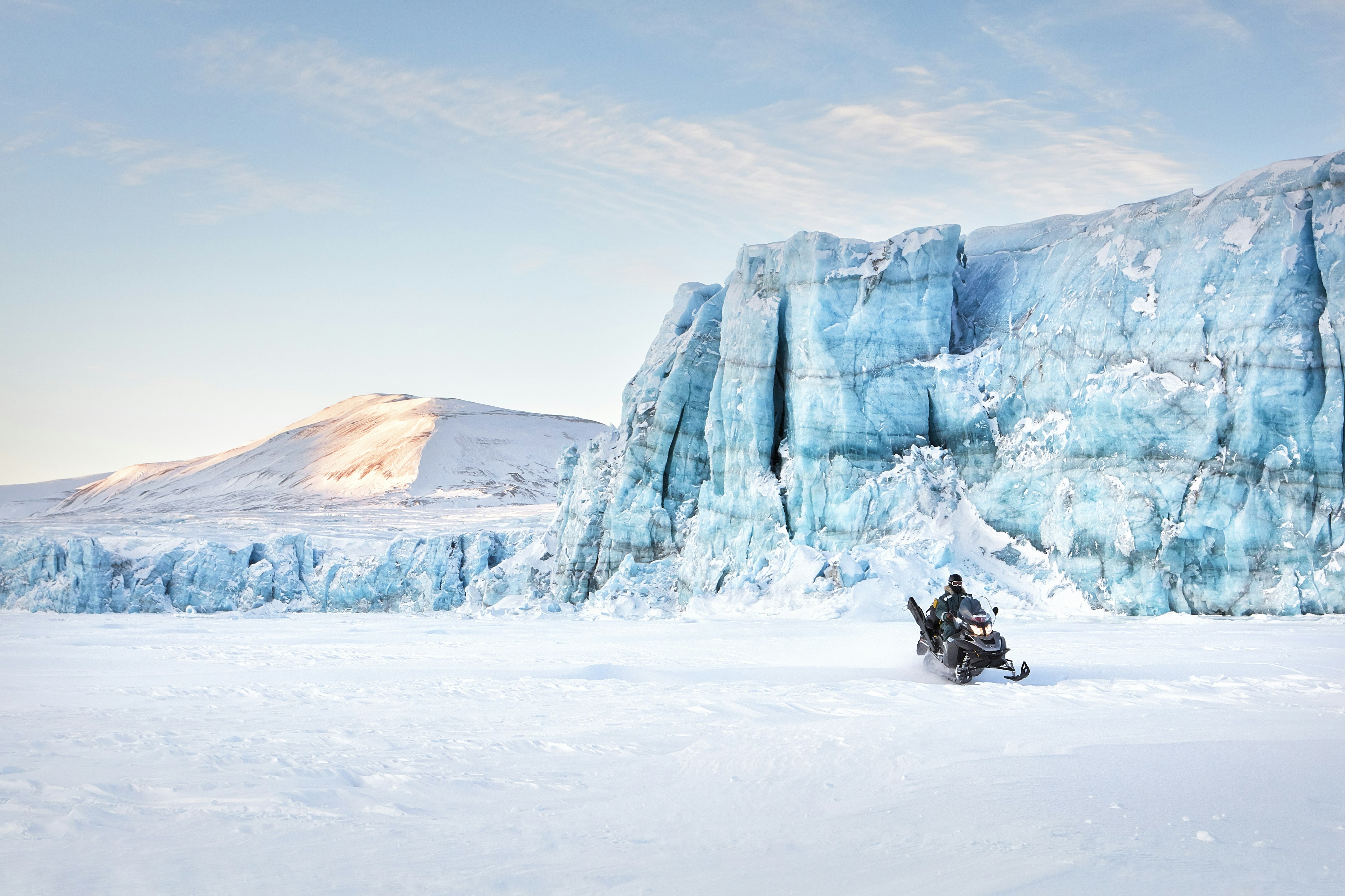 A snowmobile on frozen waters passes a massive blue-white frozen cliff face