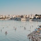 Beach and sea life in "Riviera degli Haethei" beach. 
This beach is called by the locals Spiaggia della Madonna dell'Alto Mare, taking its name from the small church that is located on top of the beach dedicated to Madonna dell'Alto Mare, one of the protectors of the city.
This beach is characterized by crystal clear water and shallow water where you can walk for a long time before reaching the high water. For this reason it is the favorite destination of families with small children, who can swim and play on the shore freely.
On the other side you can see the pier and the port as well as the center of Otranto and its old town.
August 2024: Lecce, Puglia, Italy