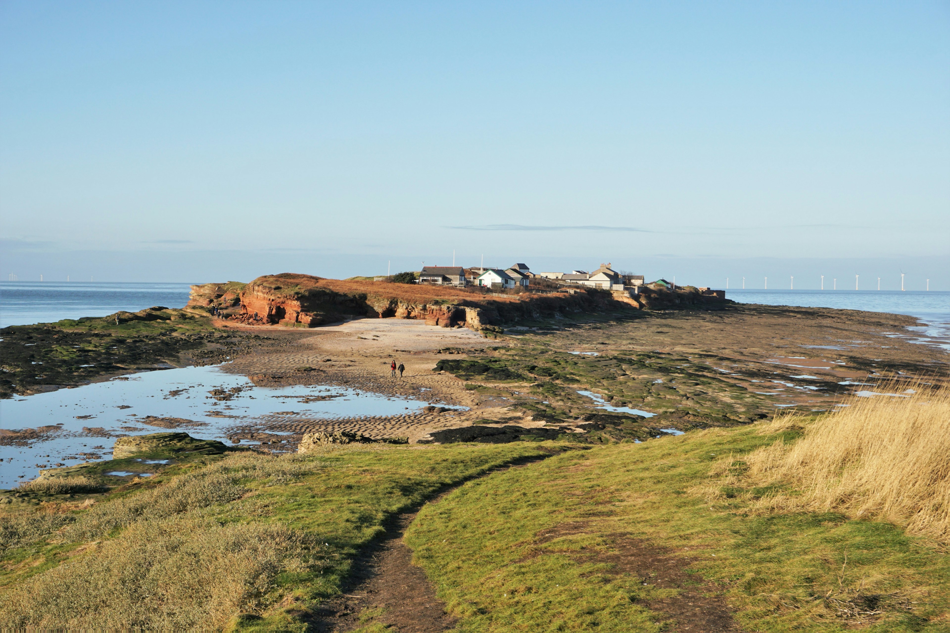 Looking towards Hilbre Island from Little Hilbre Island, located in the estuary of the River Dee. At low tide people can walk from West Kirby to the Hilbre Islands.  Two walkers are walking along the exposed river bed.