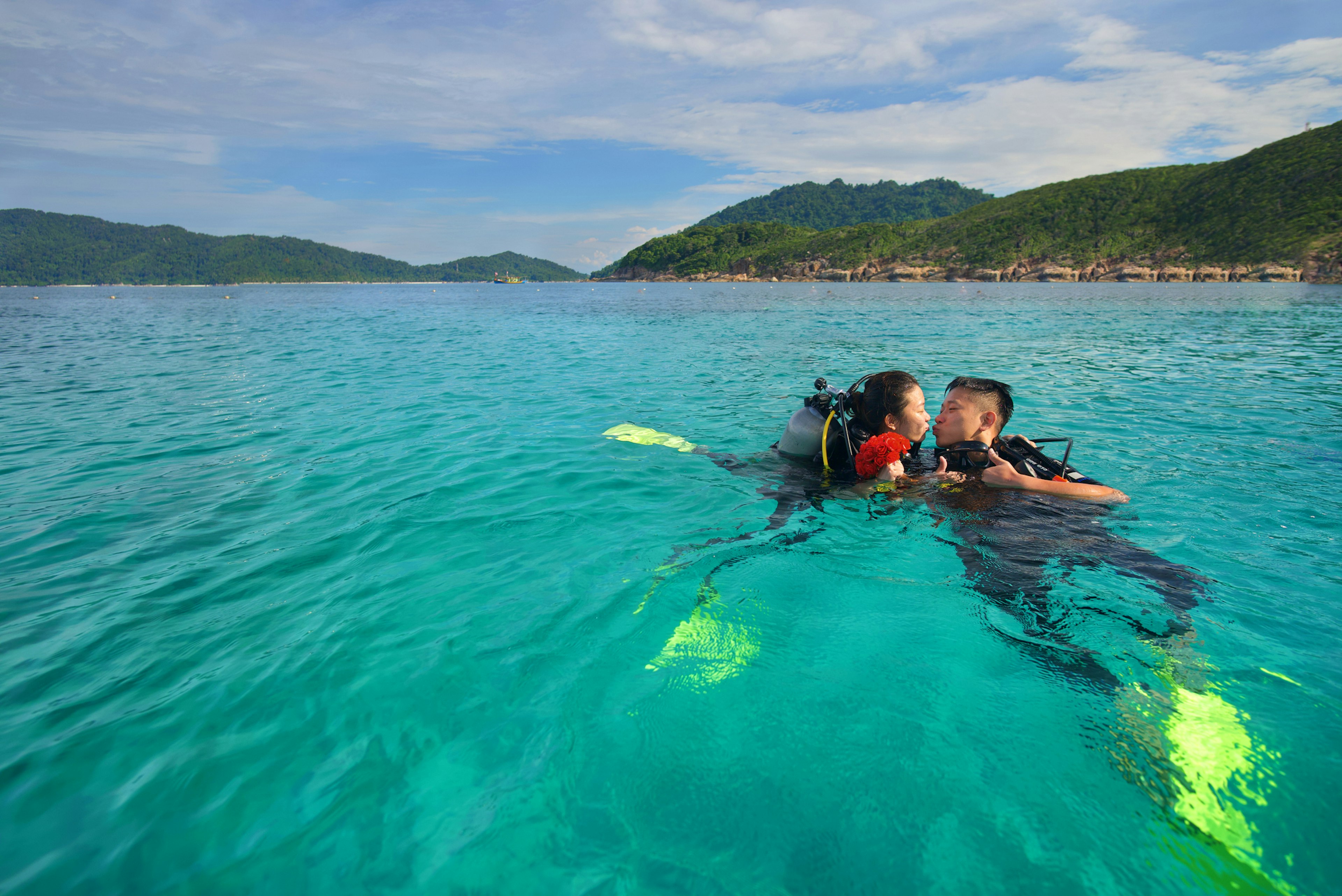 A pair of scuba divers at sea lean towards each other for a kiss on the surface in a tropical location