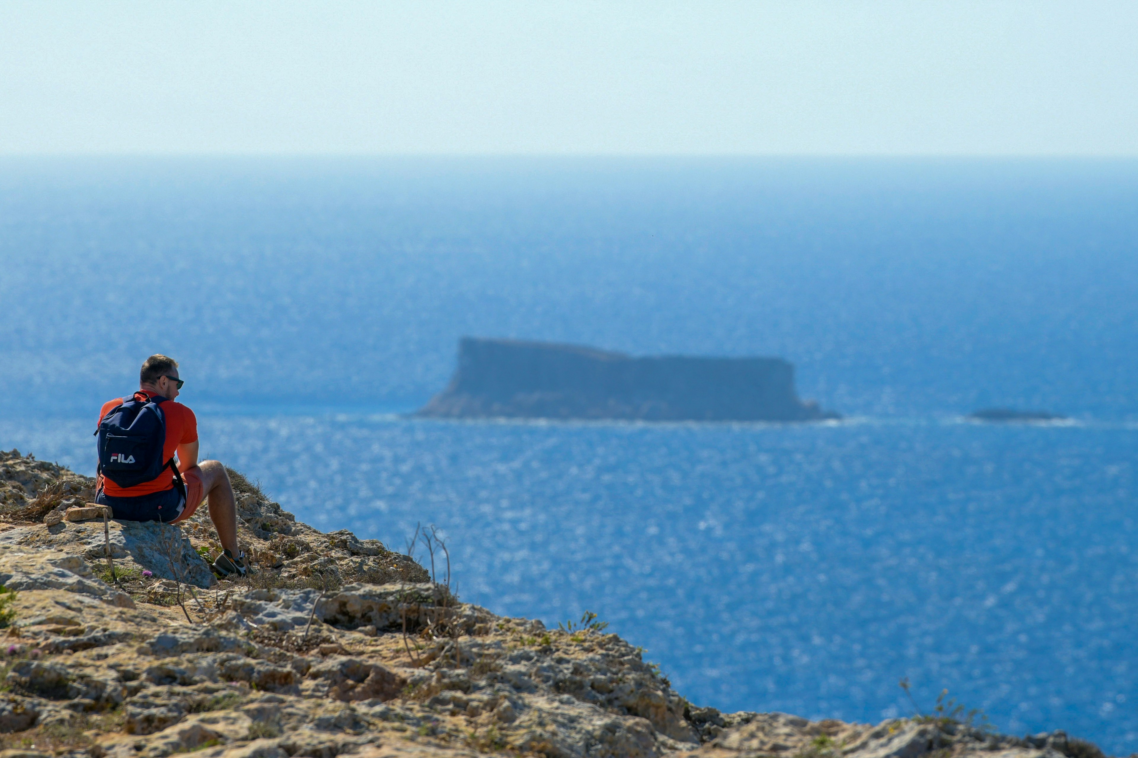 A male hiker rests on the rocky path along the Dingli Cliffs, looking out toward Filfla island, Malta