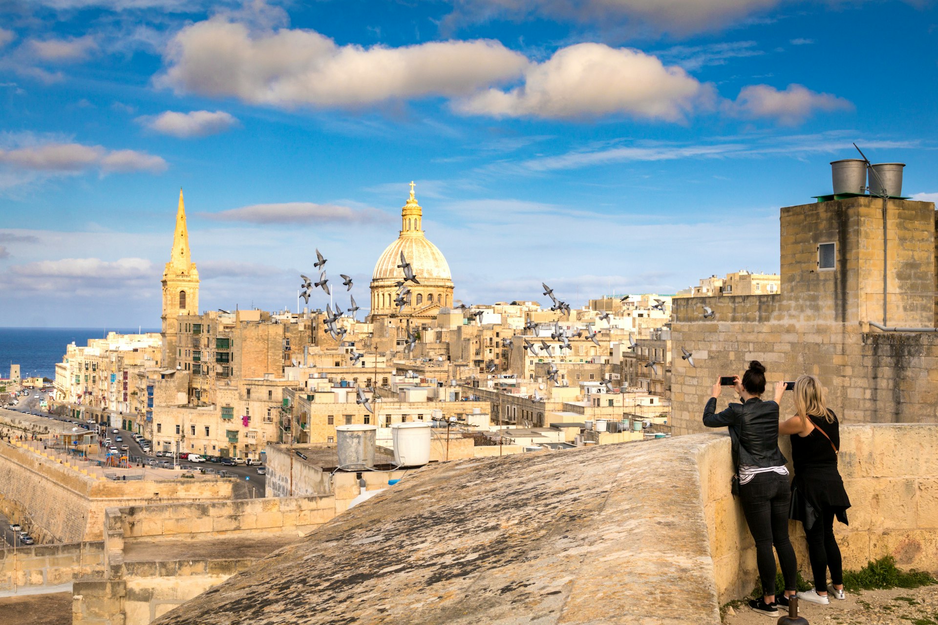 Two women tourists take photo of pigeons and beautiful skyline view of Capital city of Malta, Valletta, Dome and bell-tower above the walls, blue skies. 