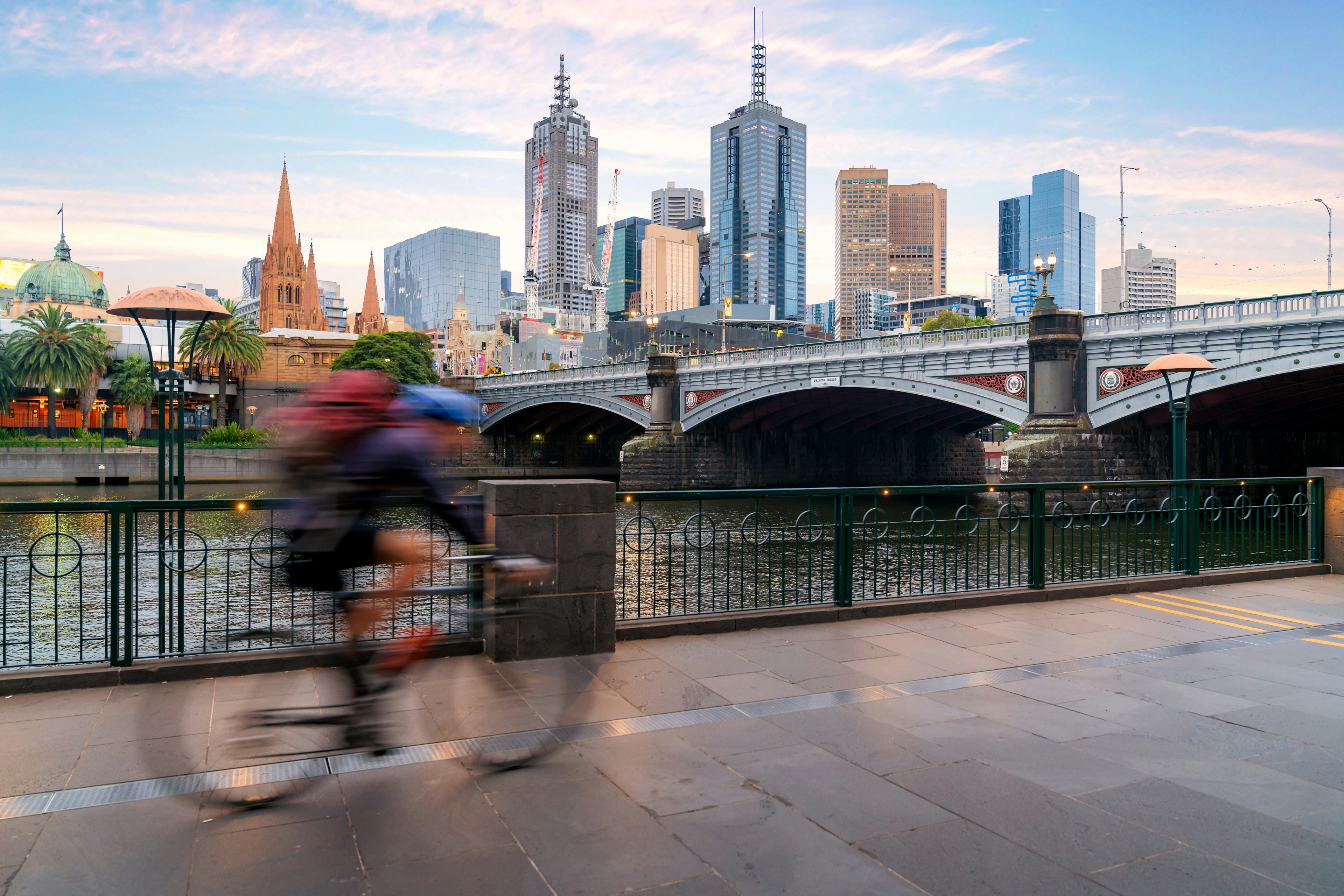 A man cycles next to the Yarra River in Melbourne, Australia. Across the river, Melbourne's main skyline is visible, with a number of towering skyscrapers.