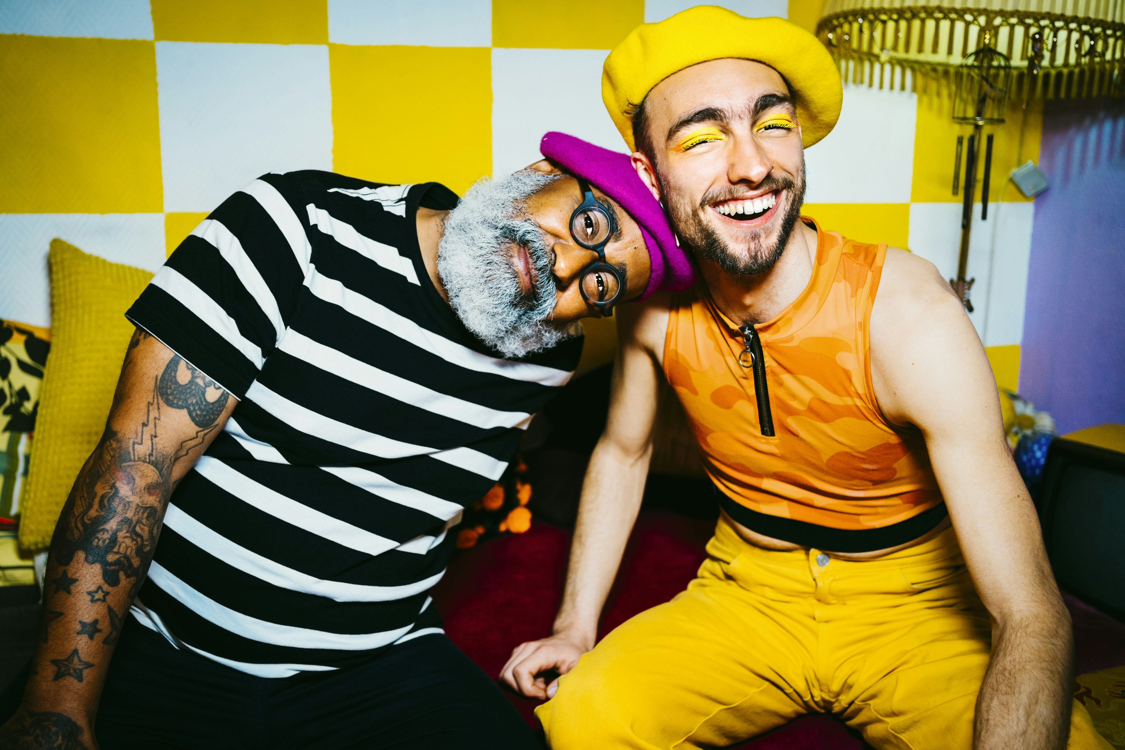 A pair of men, one older and one younger pose in front in the camera. The older male is resting his head on the younger man's shoulder while wearing a bright pink beret and a striped white and black shirt. The younger male is wearing a yellow beret and a bright orange shirt with yellow jeans.