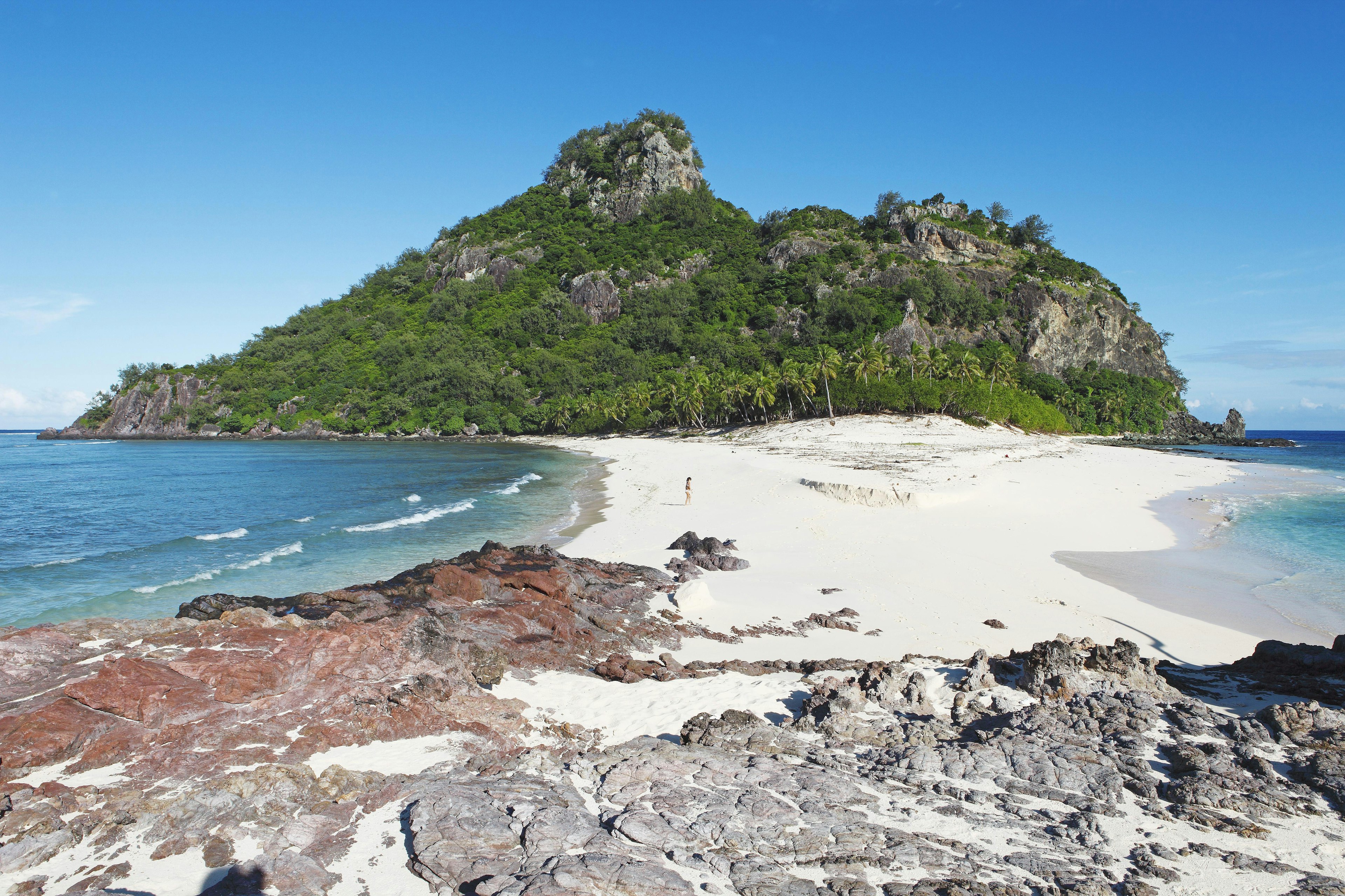 A woman walks across the sandy spit beach on Monuriki Island, Fiji as the limestone islet climbs high above her with trees clinging on