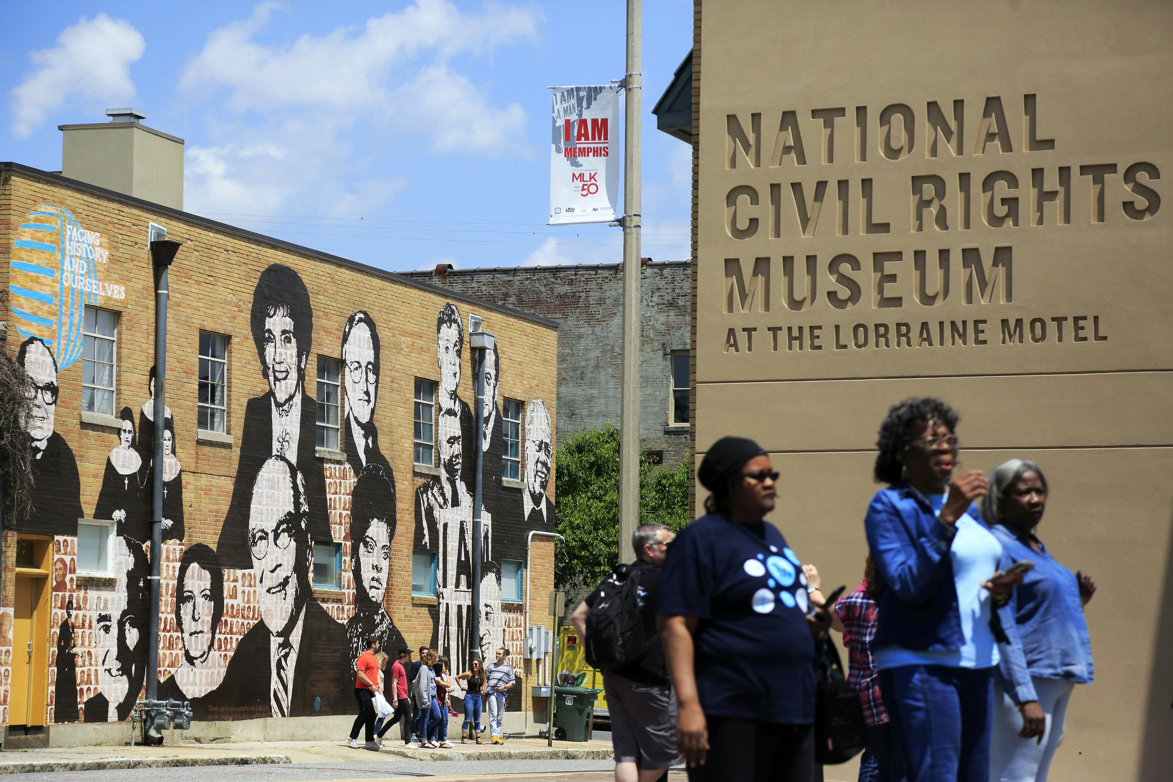 A trio of women walk towards the National Civil Rights Museum at the Lorraine Motel. In the background you can see a brick building covered in paintings of prominent Americans.