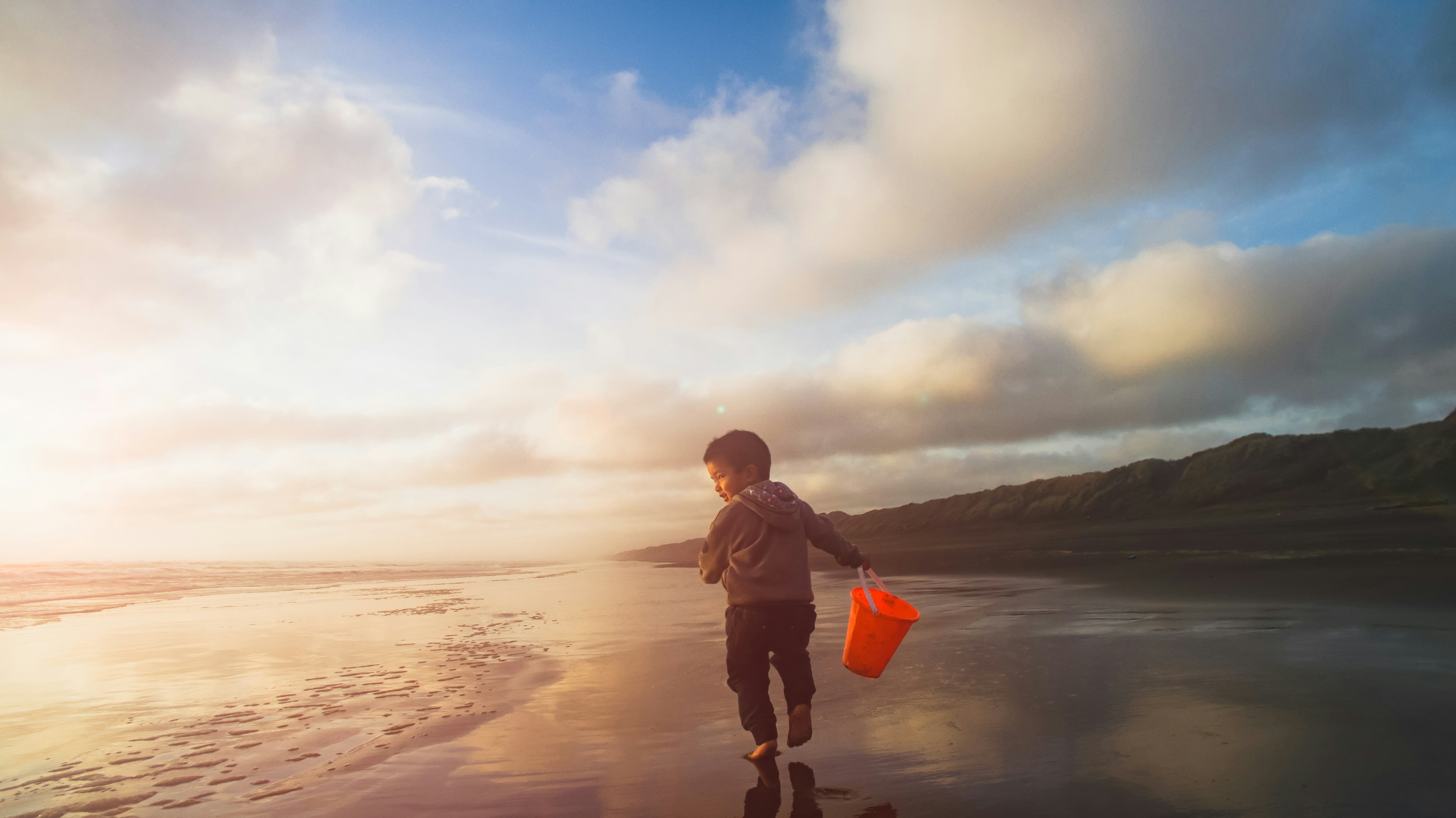 A young child runs a long an empty beach near Auckland holding a red bucket at sunset