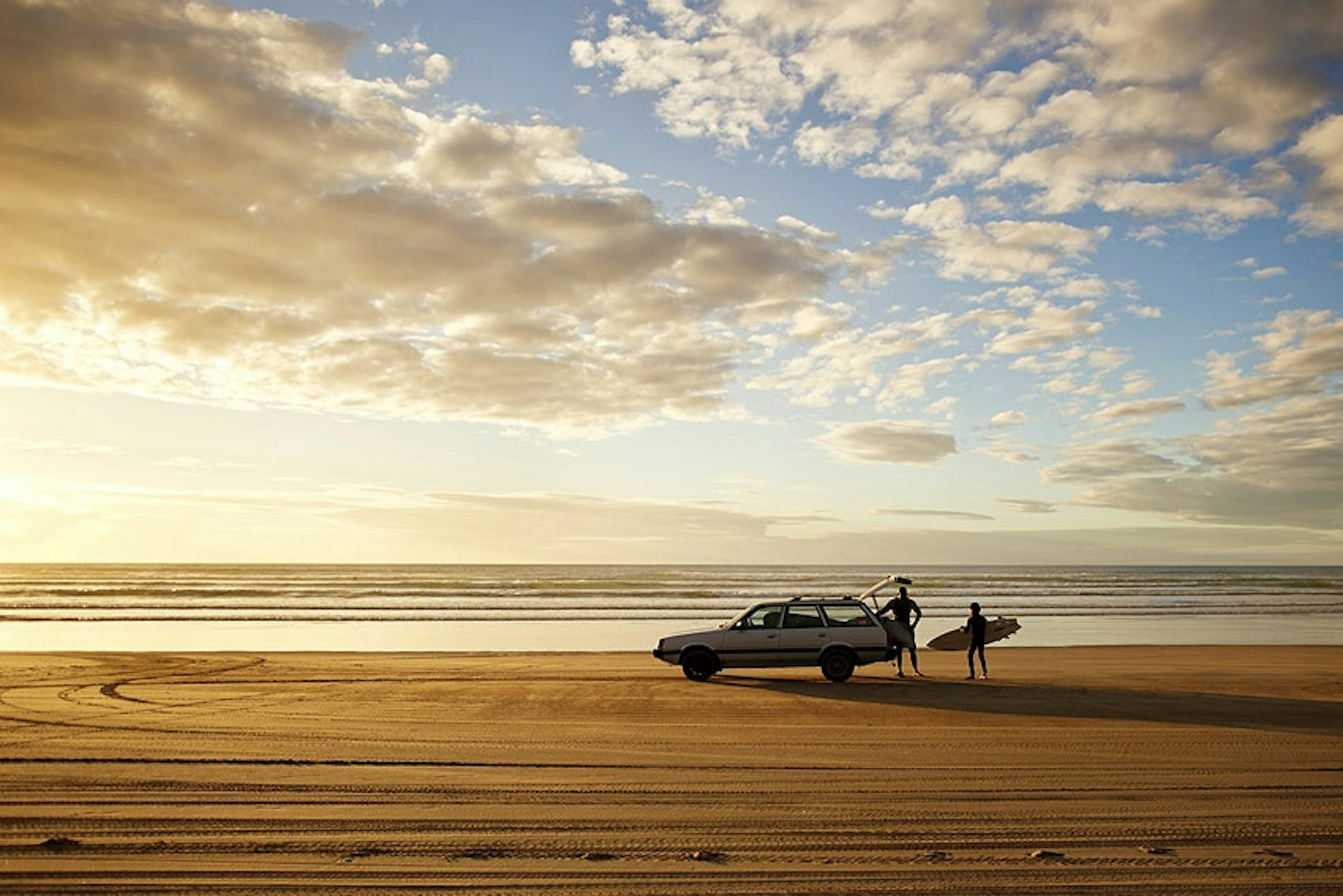 A car parked on a sandy beach as two surfers unload their boards