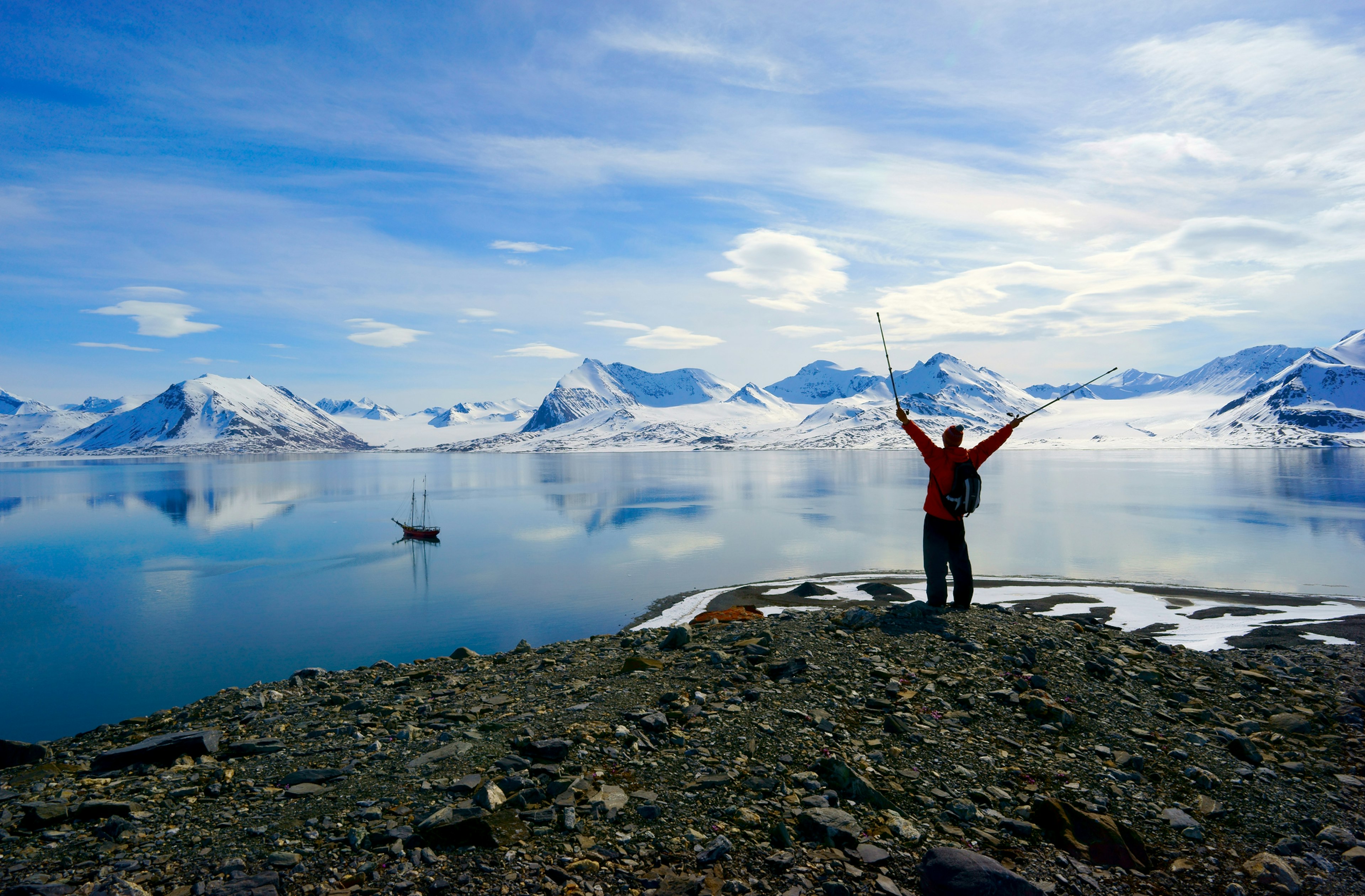 A hiker holds walking poles aloft on a clifftop at the edge of a fjord