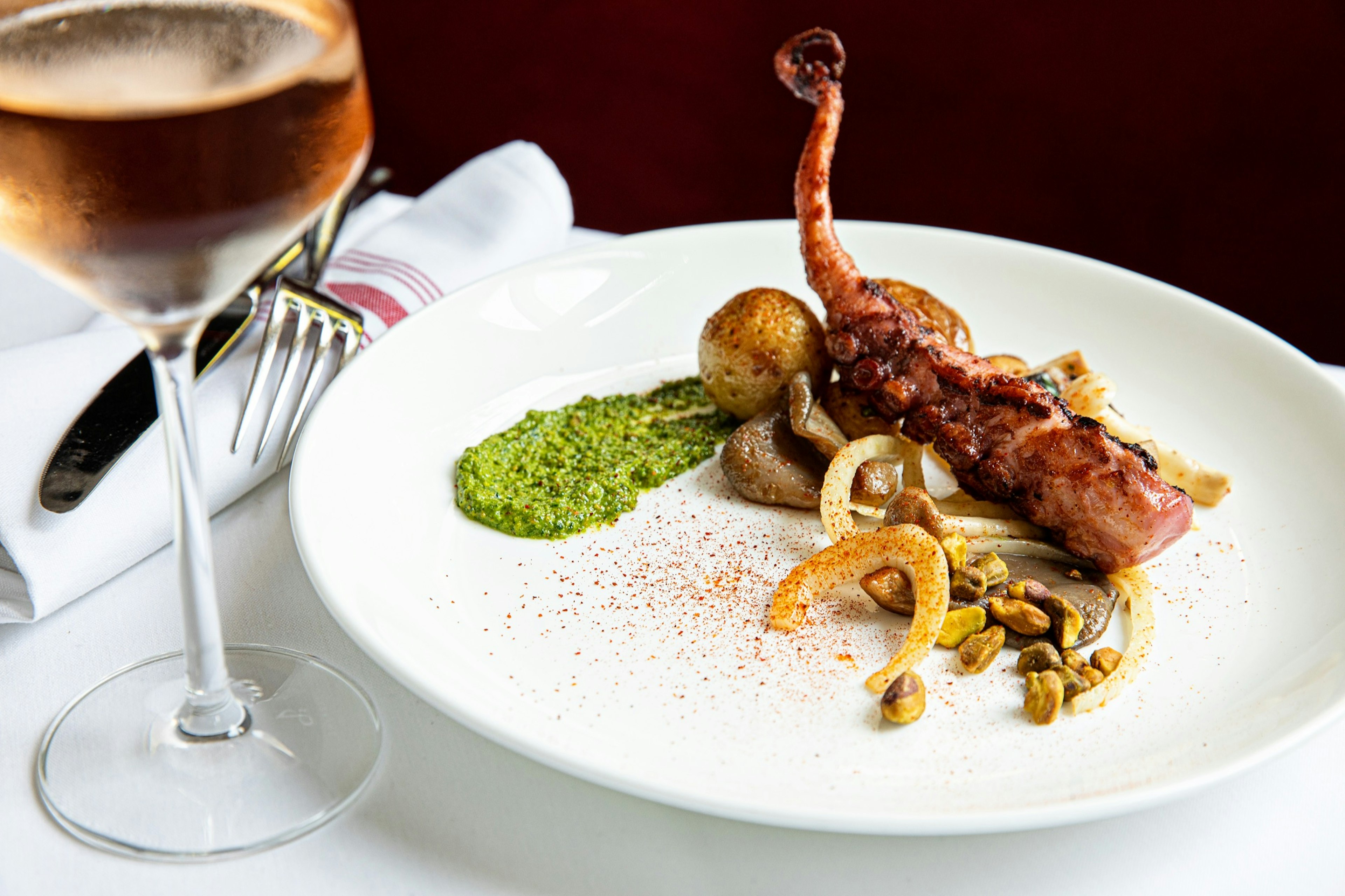 Octopus with a side of small portions of potatoes, onions and corn. There's a smear of green sauce on the side of the plate. There's a glass of wine, a fork and knife on a white tablecloth.