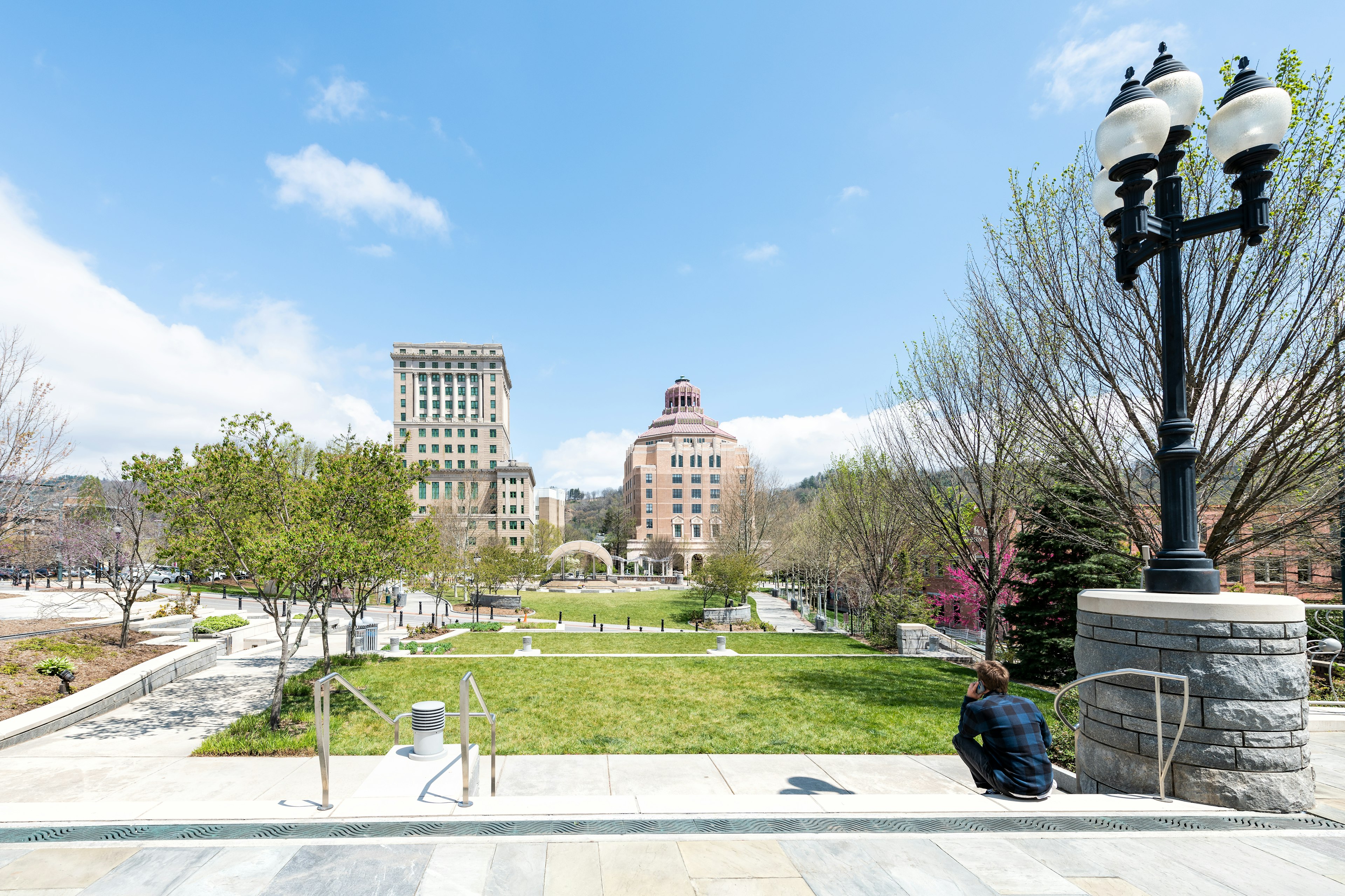 Back of young hipster millennial man sitting looking at buildings, courthouse and green Pack Square Park in hippie North Carolina NC famous town, city in the mountains, talking on phone