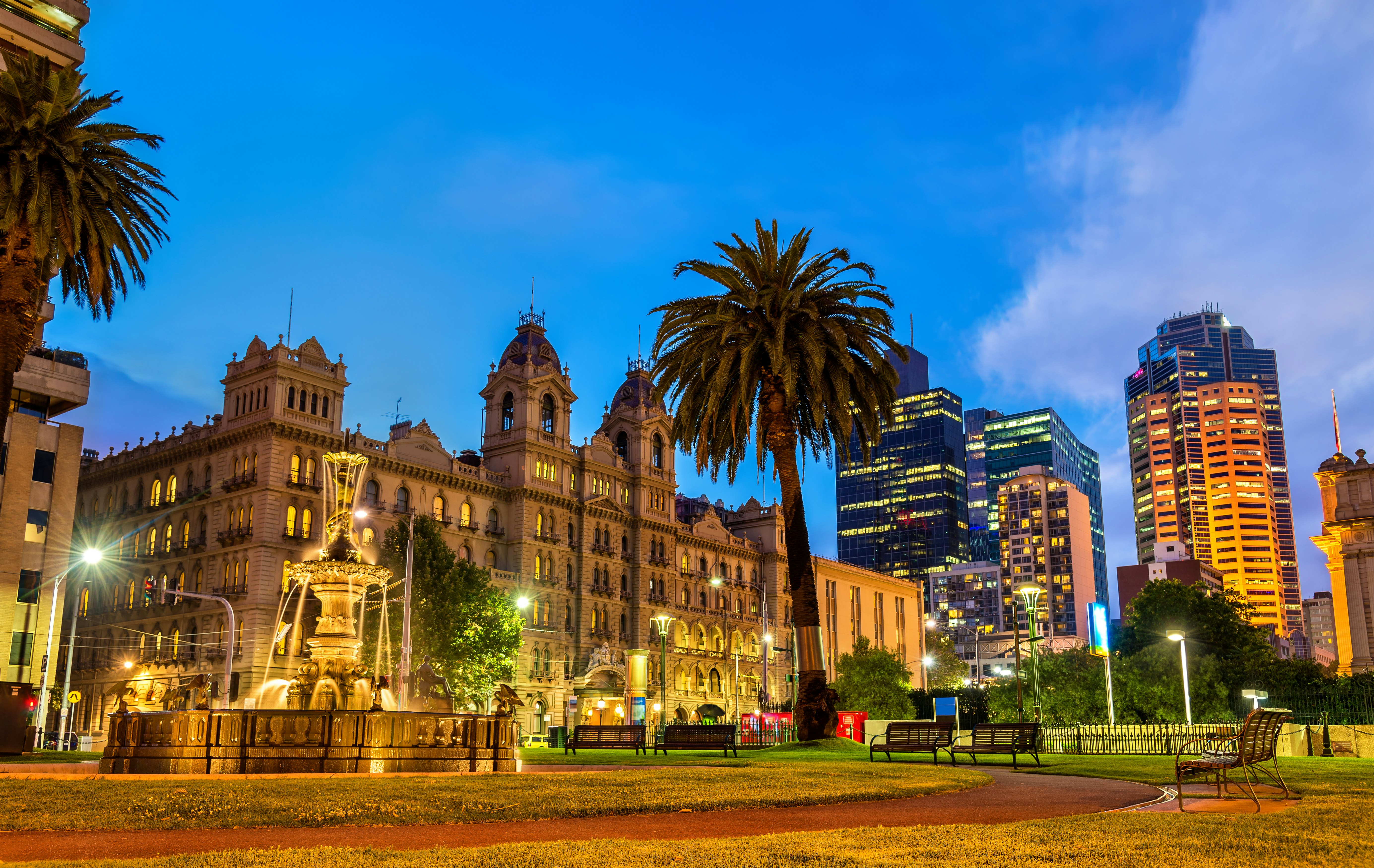 A view of Parliament House, and the adjacent garden, in Melbourne, Australia. The building is large and grand, and is lit up in the dark of the evening.