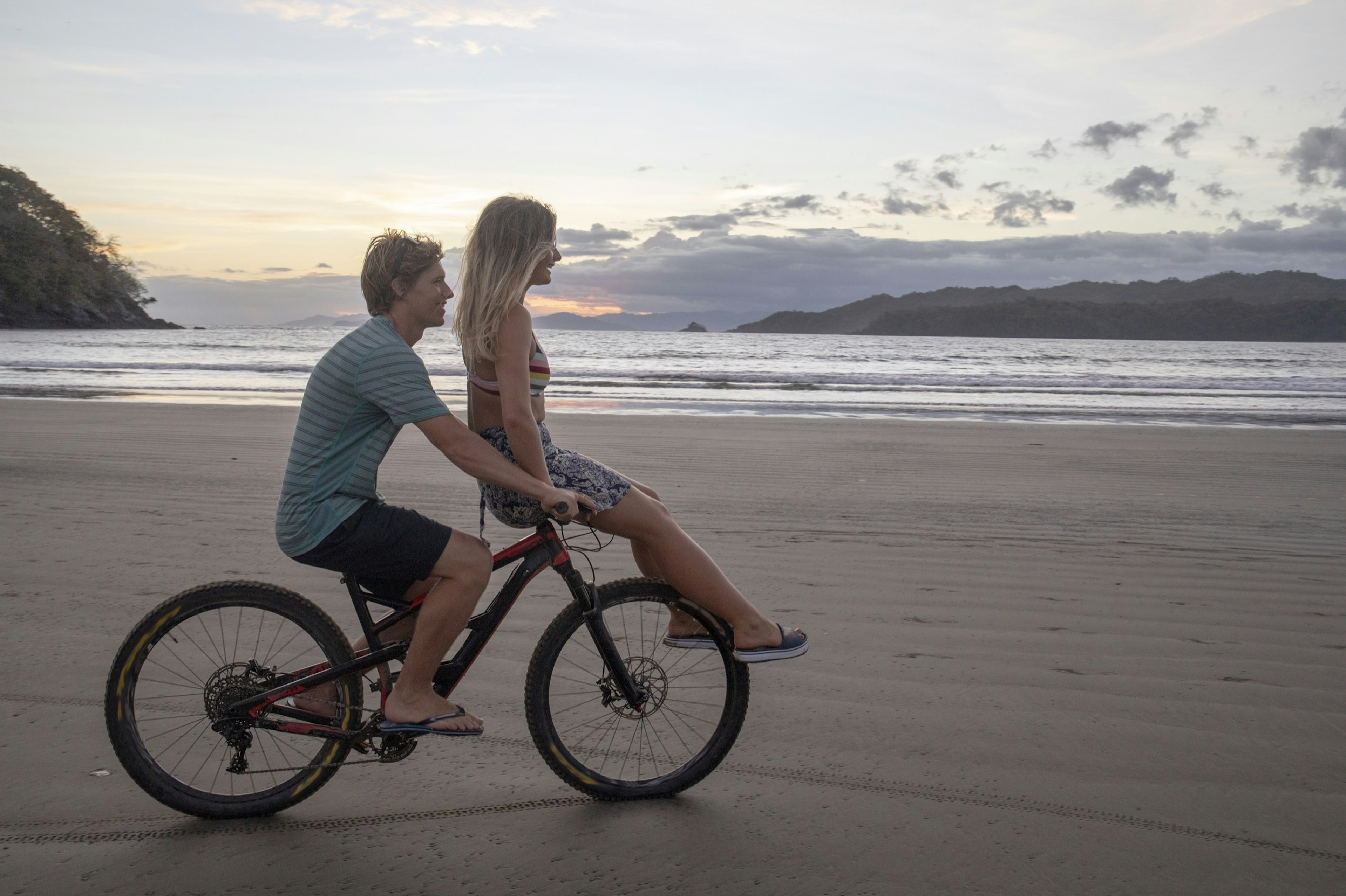 A man steers a bike while a young woman sits on top of the handlebars as they bike on Playa Venao in Panama.