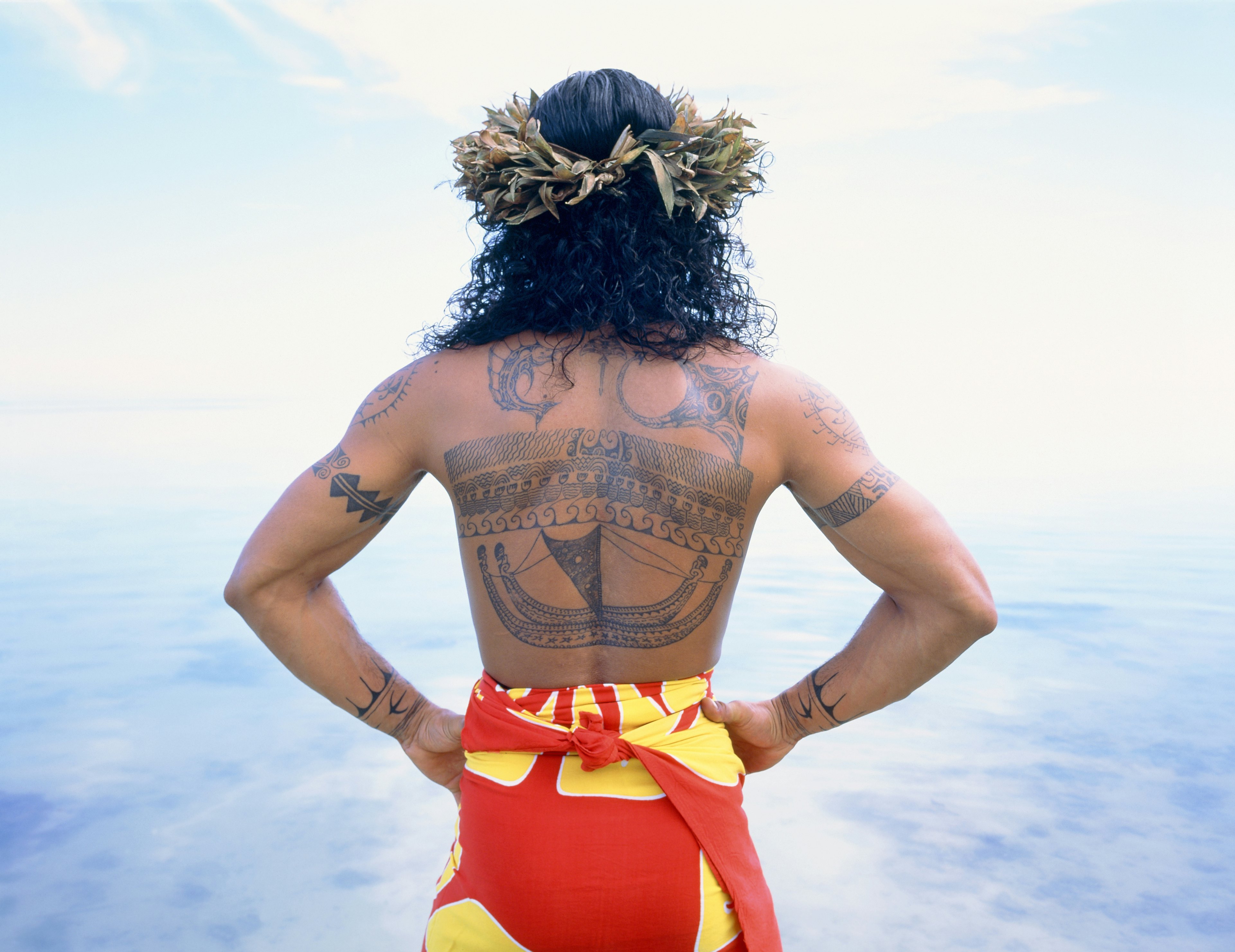 A man with long curly hair wears a floral headdress and a bright yellow and red sarong. His back is covered with traditional Polynesian tattoos.