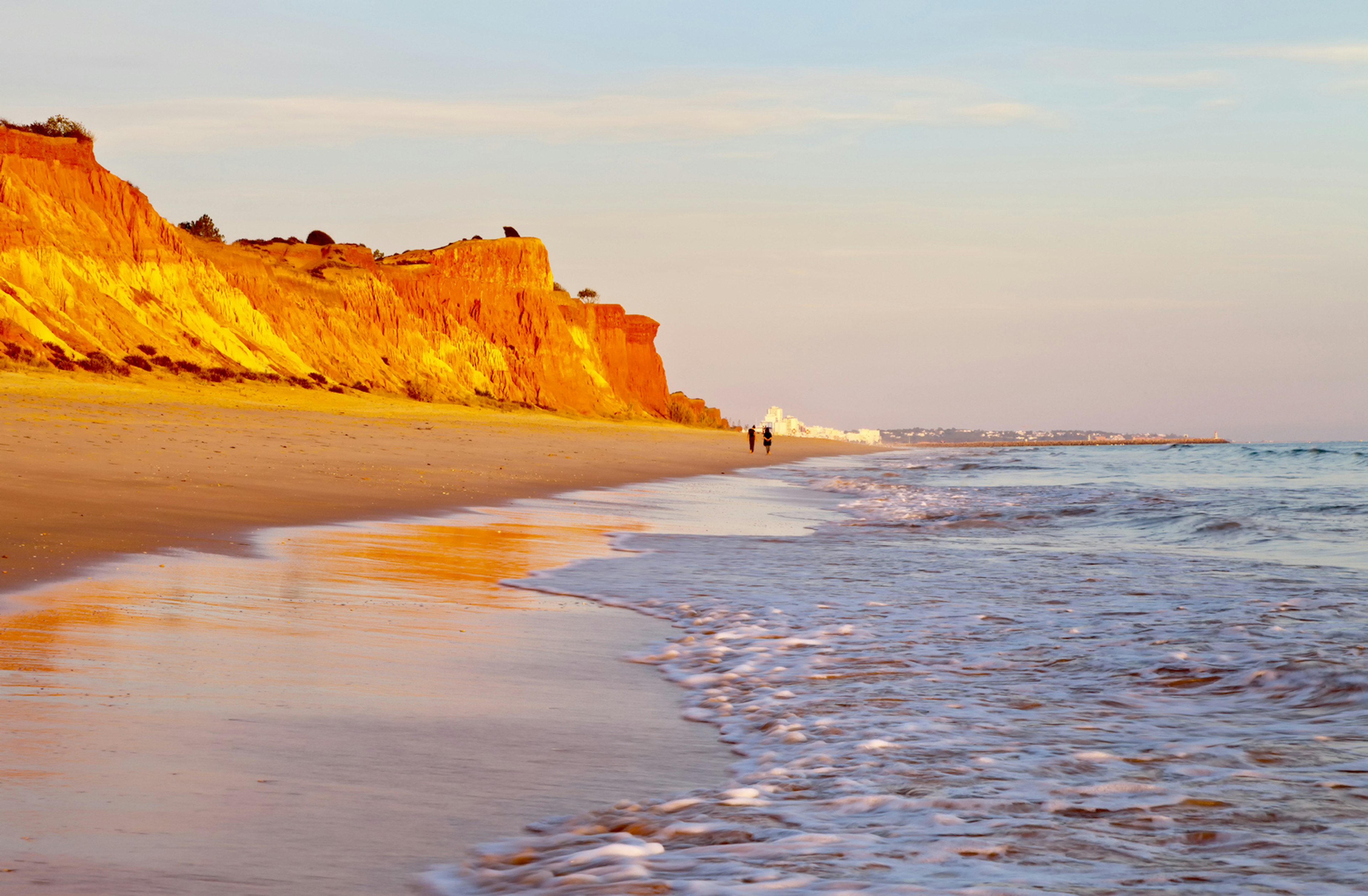 A couple in silhouette walk along a beach backed by cliffs that have turned red in the sunlight as it sets