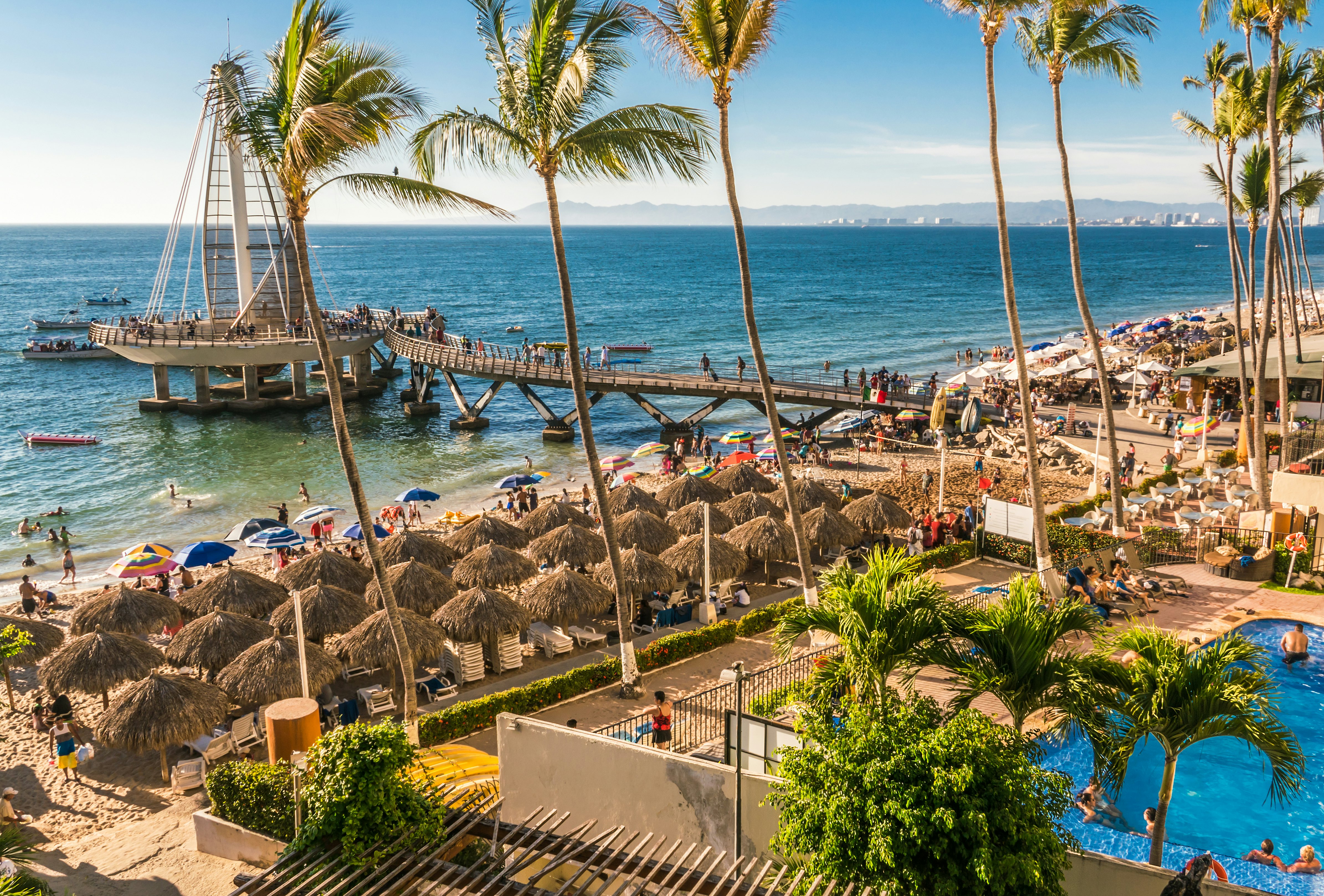 A crowded beach scene with people walking the seafront or relaxing under palm-frond shades in Puerto Vallarta.