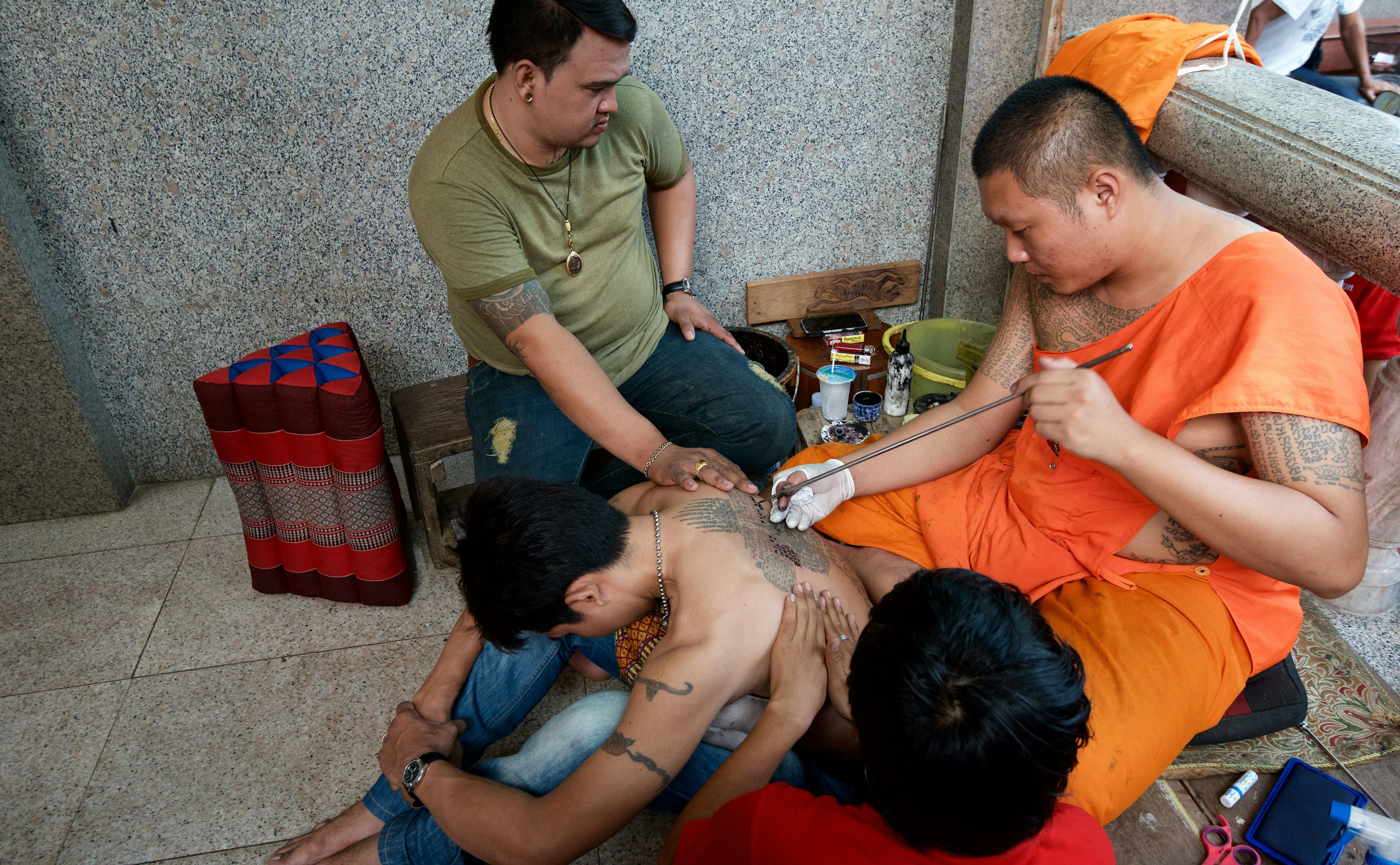 A man bends forward as his back is being tattooed by a Buddhist monk. He is being held by two men.