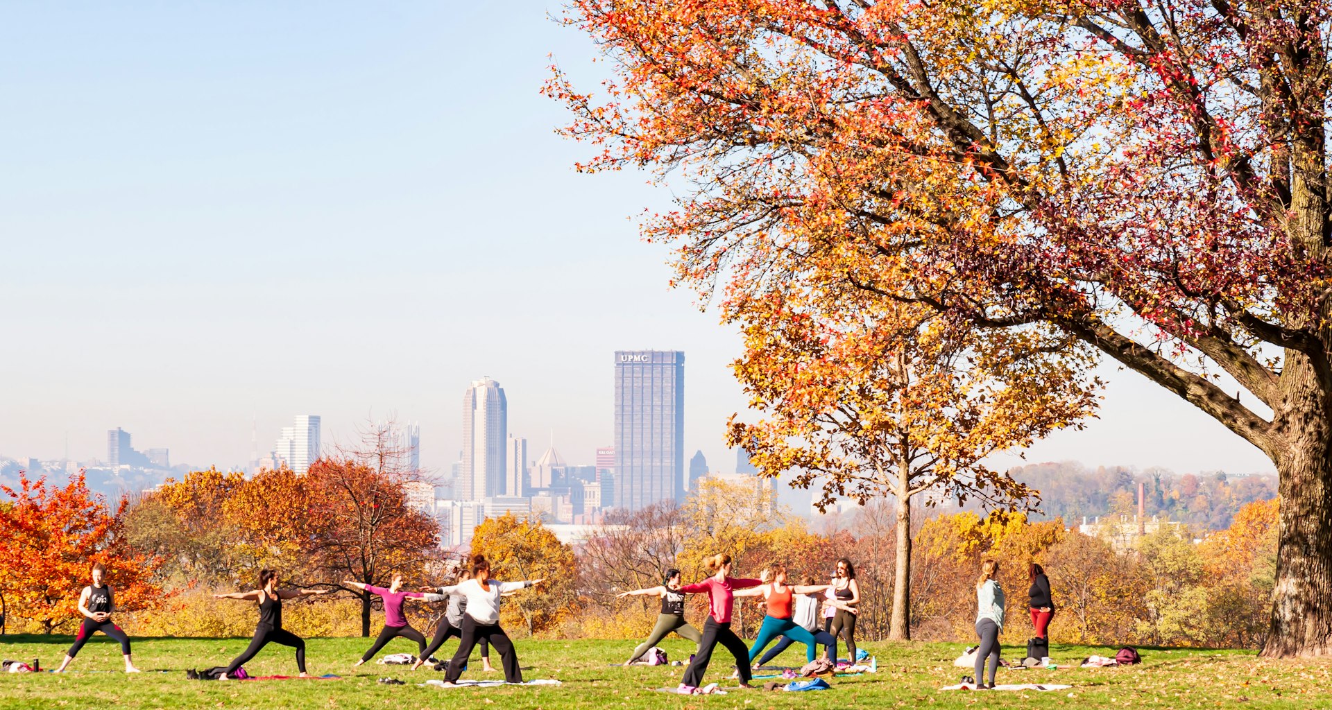 A group of women having a yoga class in Schenley Park with downtown Pittsburgh in the background on a hazy fall morning