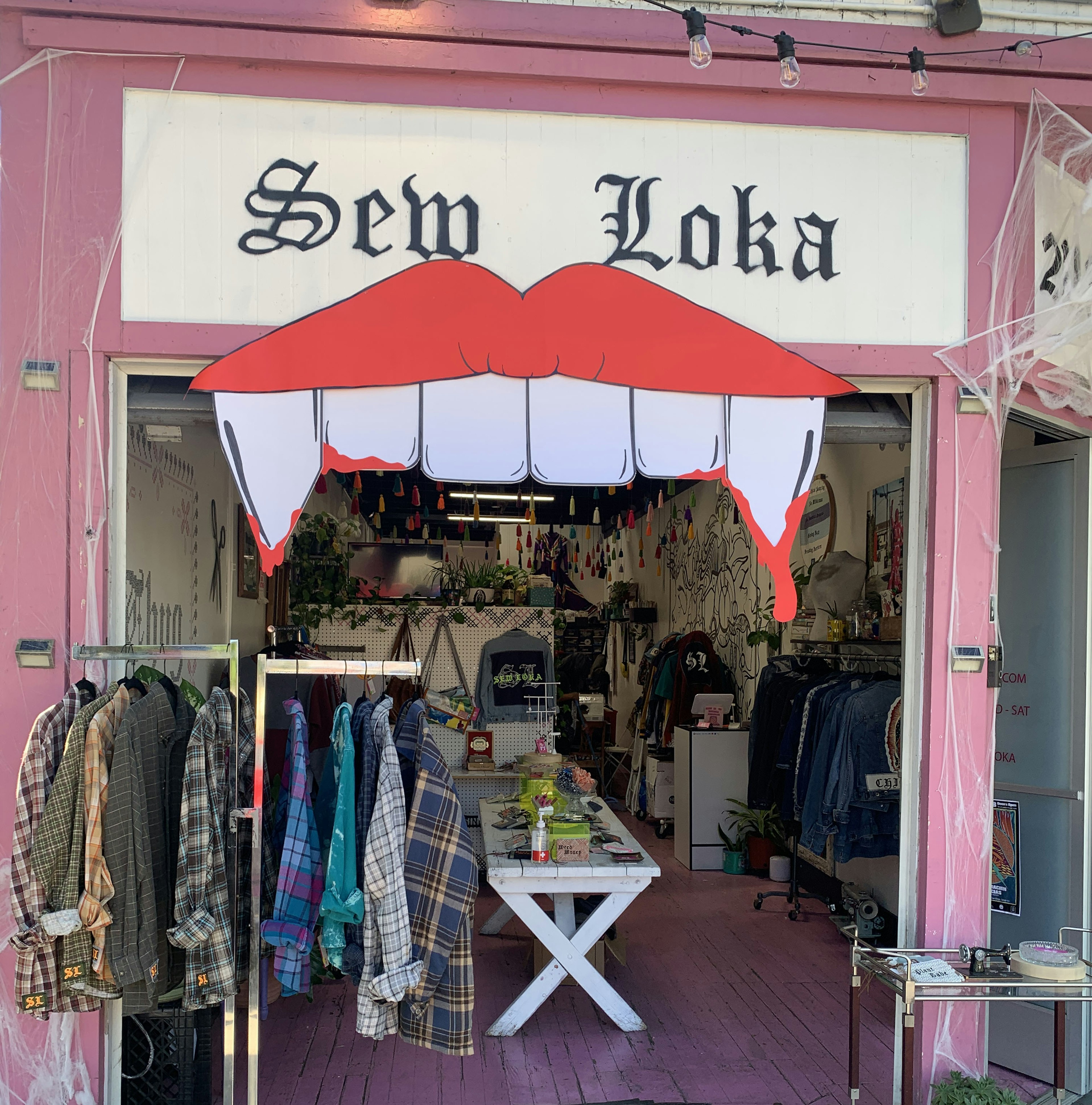 Exterior shot of the Sew Loka shop. There is a sign in the shape of a pair of lips with fangs hanging from the storefront. You can see a clothing rack and the interior of the store.