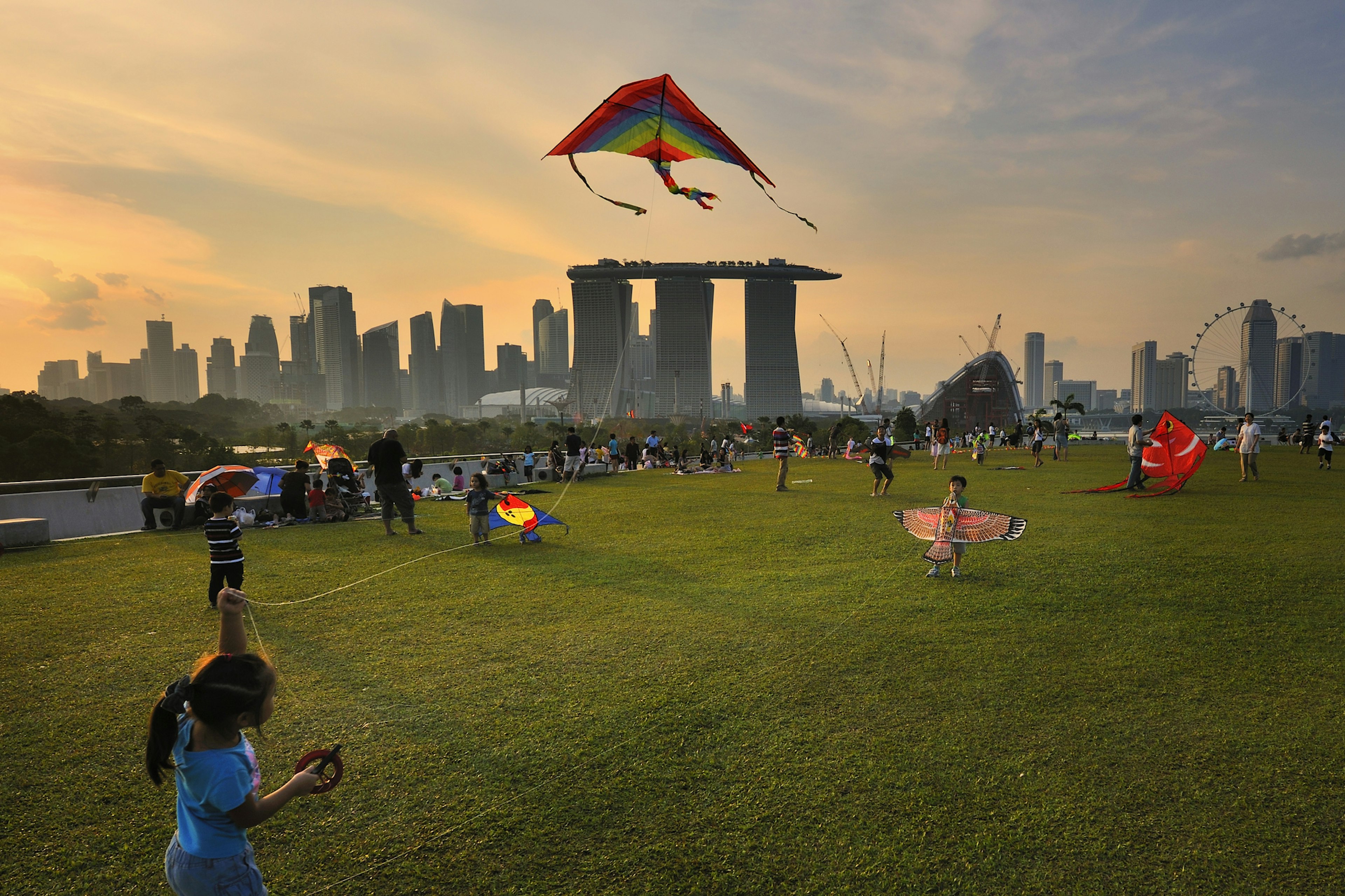 Families flying kites in a city's green space as the sun sets over the tall buildings on the skyline