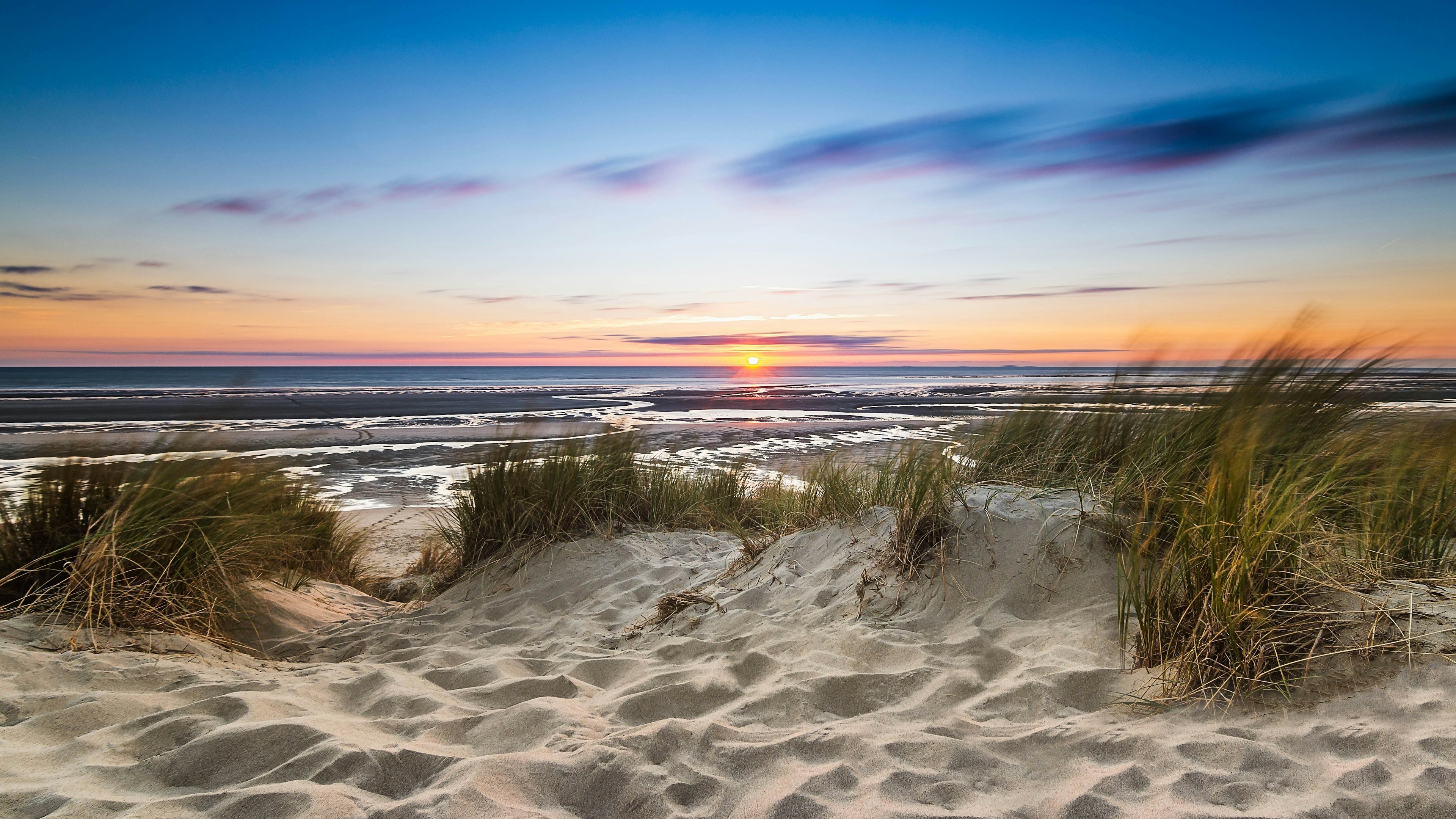 Sand dunes on the edge of a body of water with the sun setting