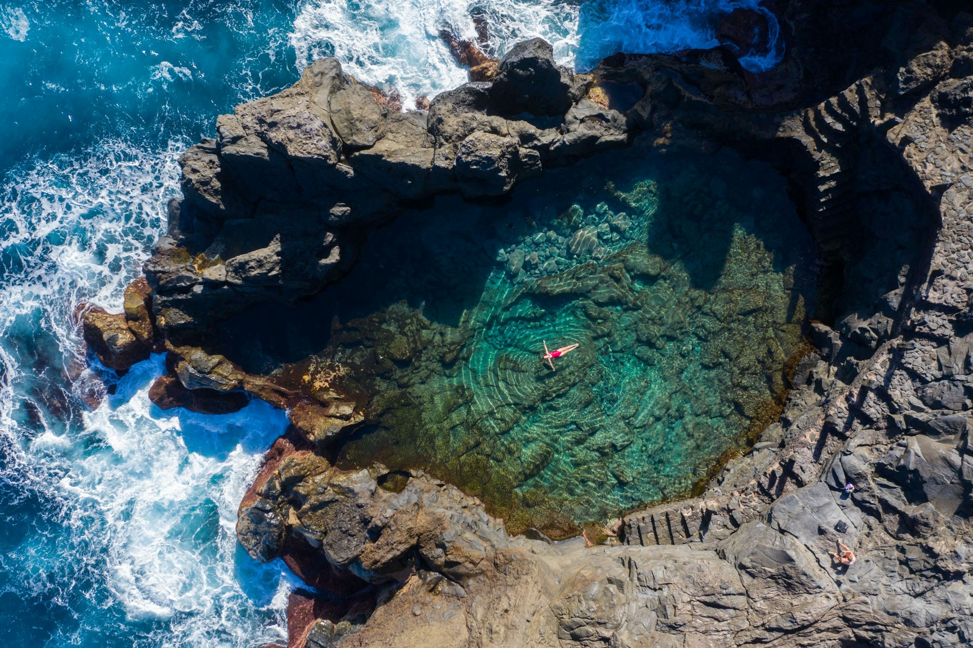 A woman in a red bathing suit floats in a large rock pool next to the crashing surf