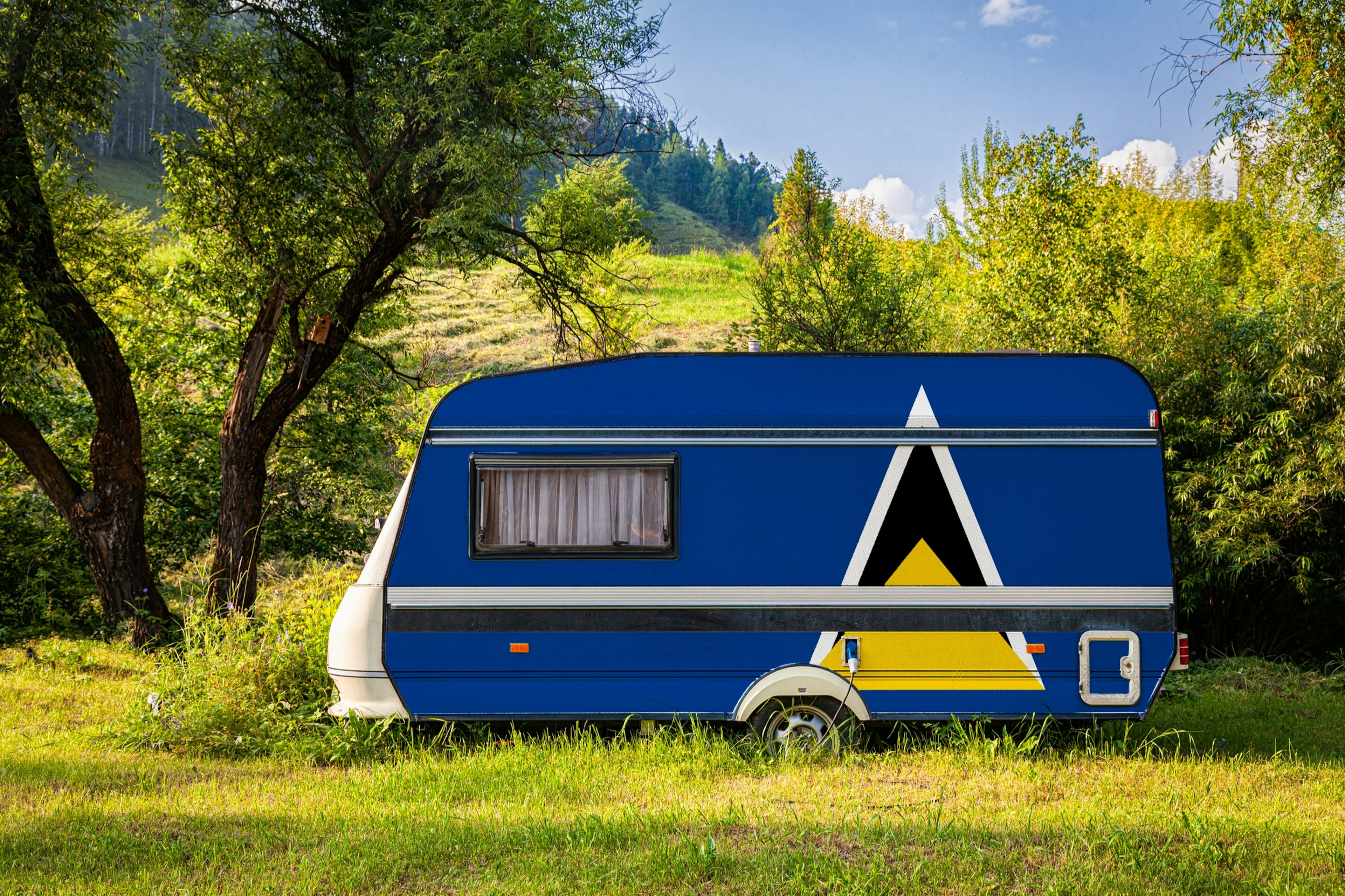 A car trailer, a motor home, painted in the national flag of Saint Lucia is parked on grass in a mountainous region