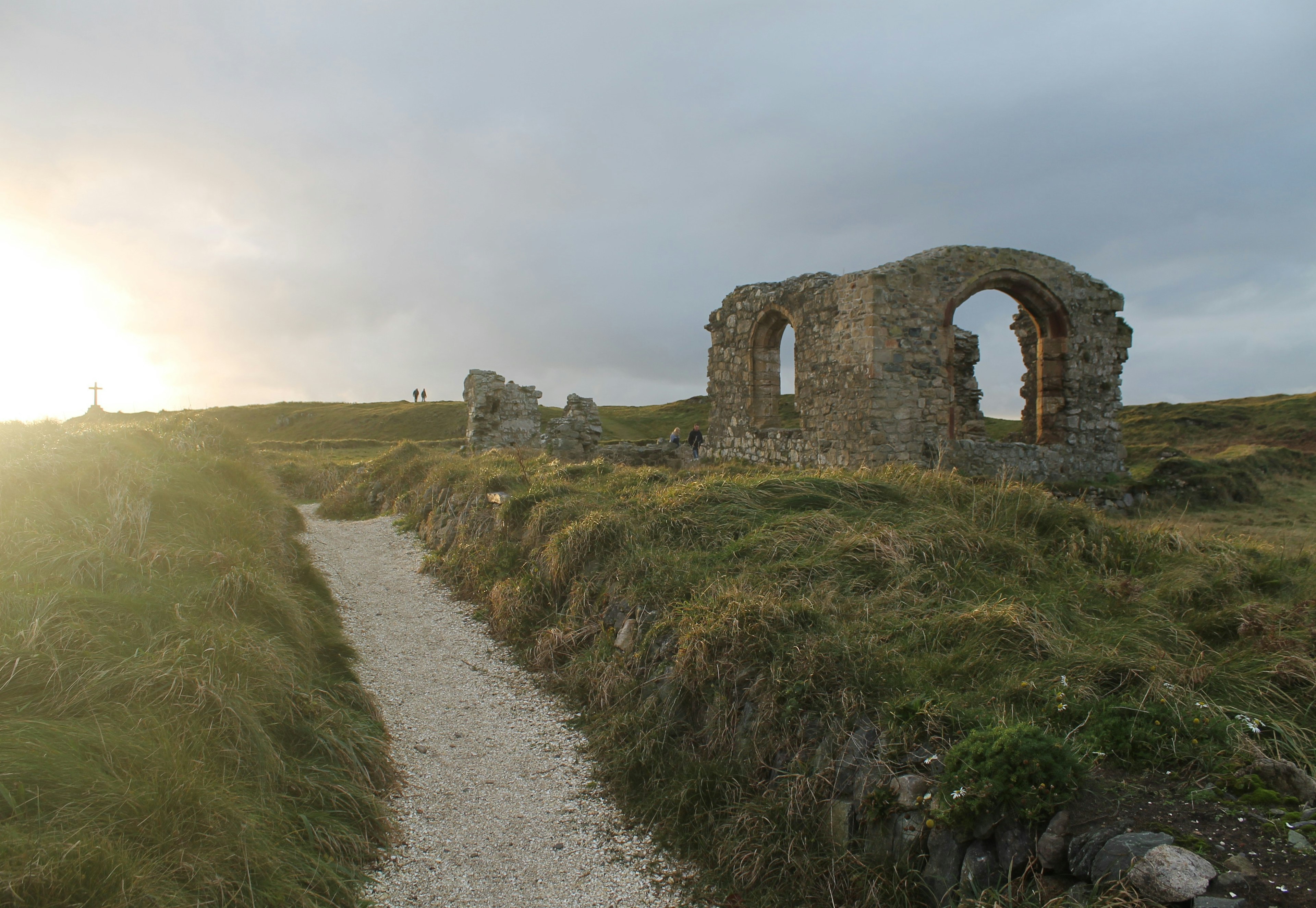 St Dwynwen's Church in Anglesey at sunset. The church is essentially a ruin, with only parts of the walls remaining. Surrounding the church is thick grassland with a narrow walkway to the entrance.