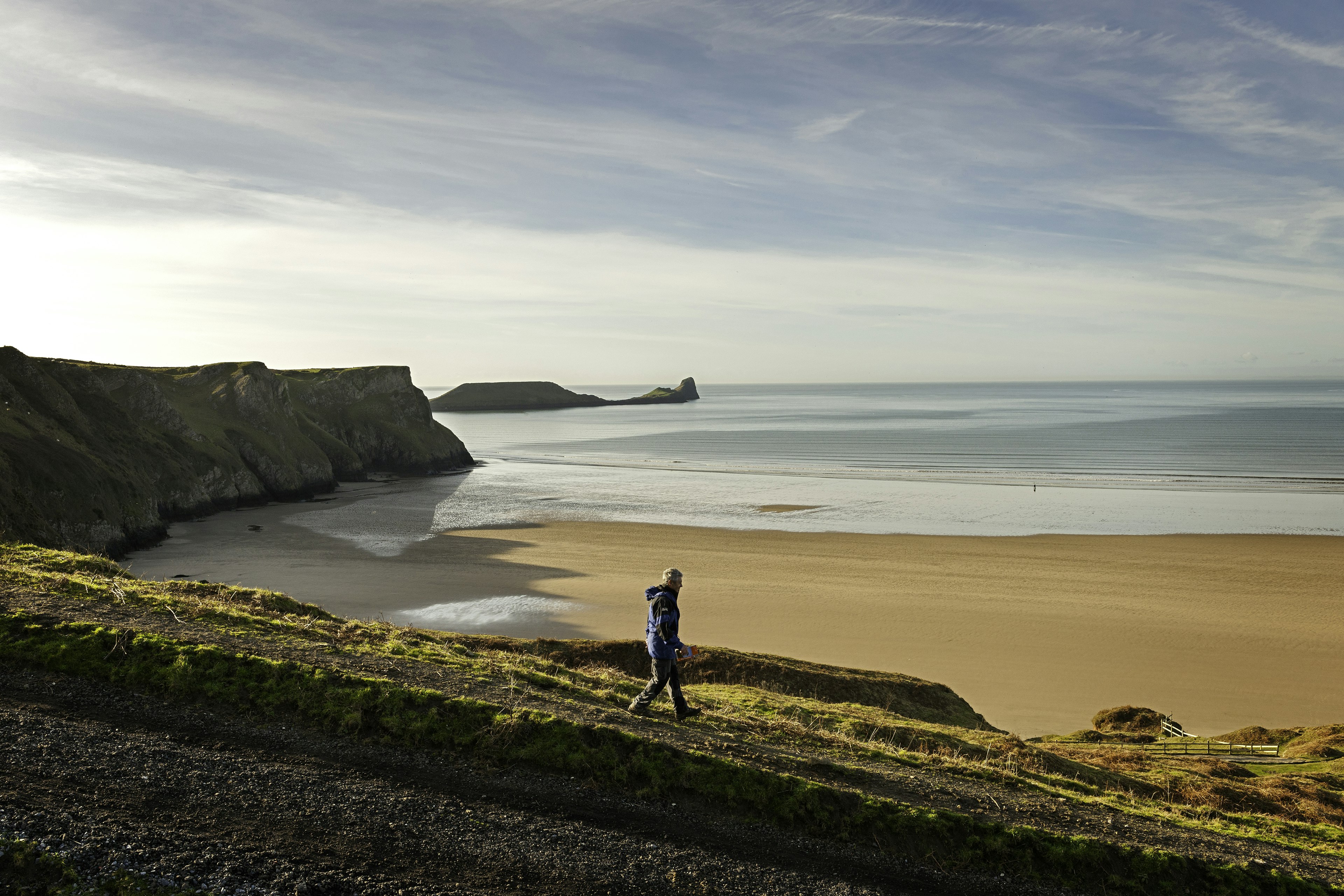 Man walking above Rhossili Beach, with Worm's Head in the background