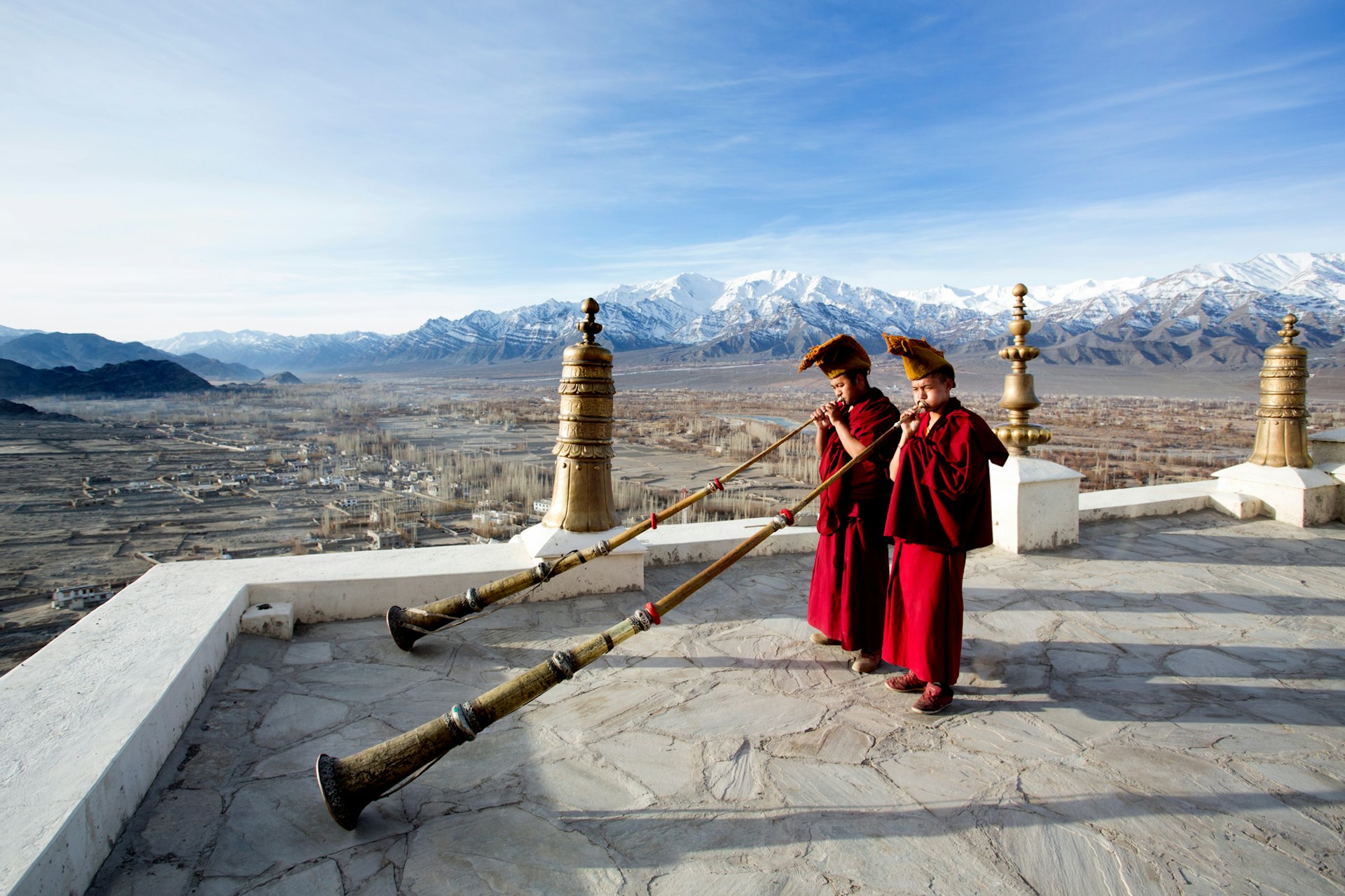 Red-robed monks blow horns on the rooftop of a Ladakh monastery with mountains beyond. 