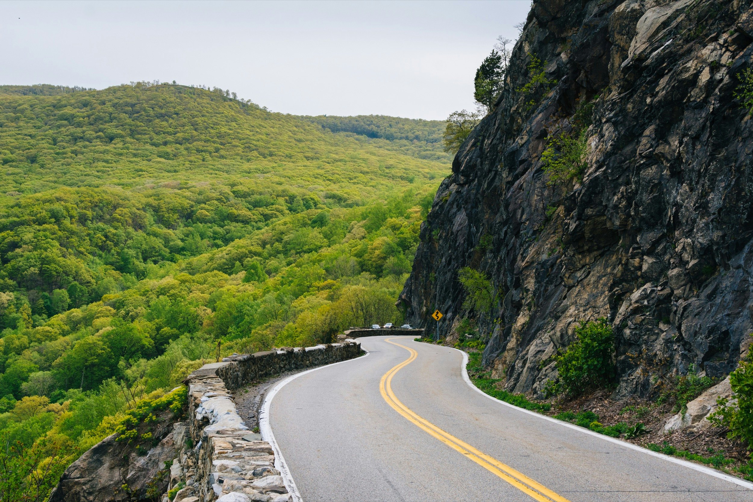 A two-lane highway in Cornwall on the Hudson, NY. On one side of the highway is a dense forested area. On the other side a tall mountain.