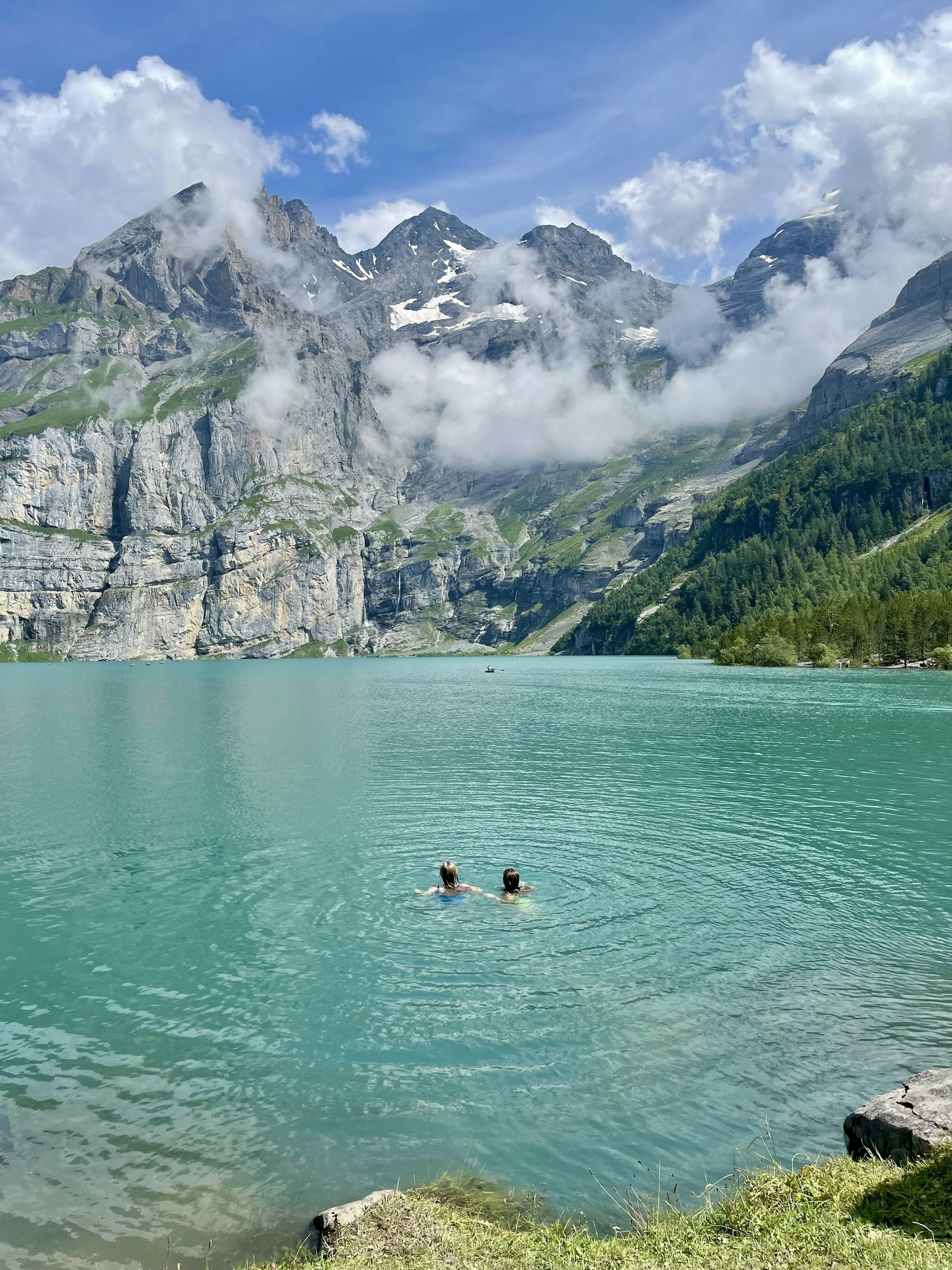 Two women swimming in turquoise waters surrounded by high mountain peaks with snow on them