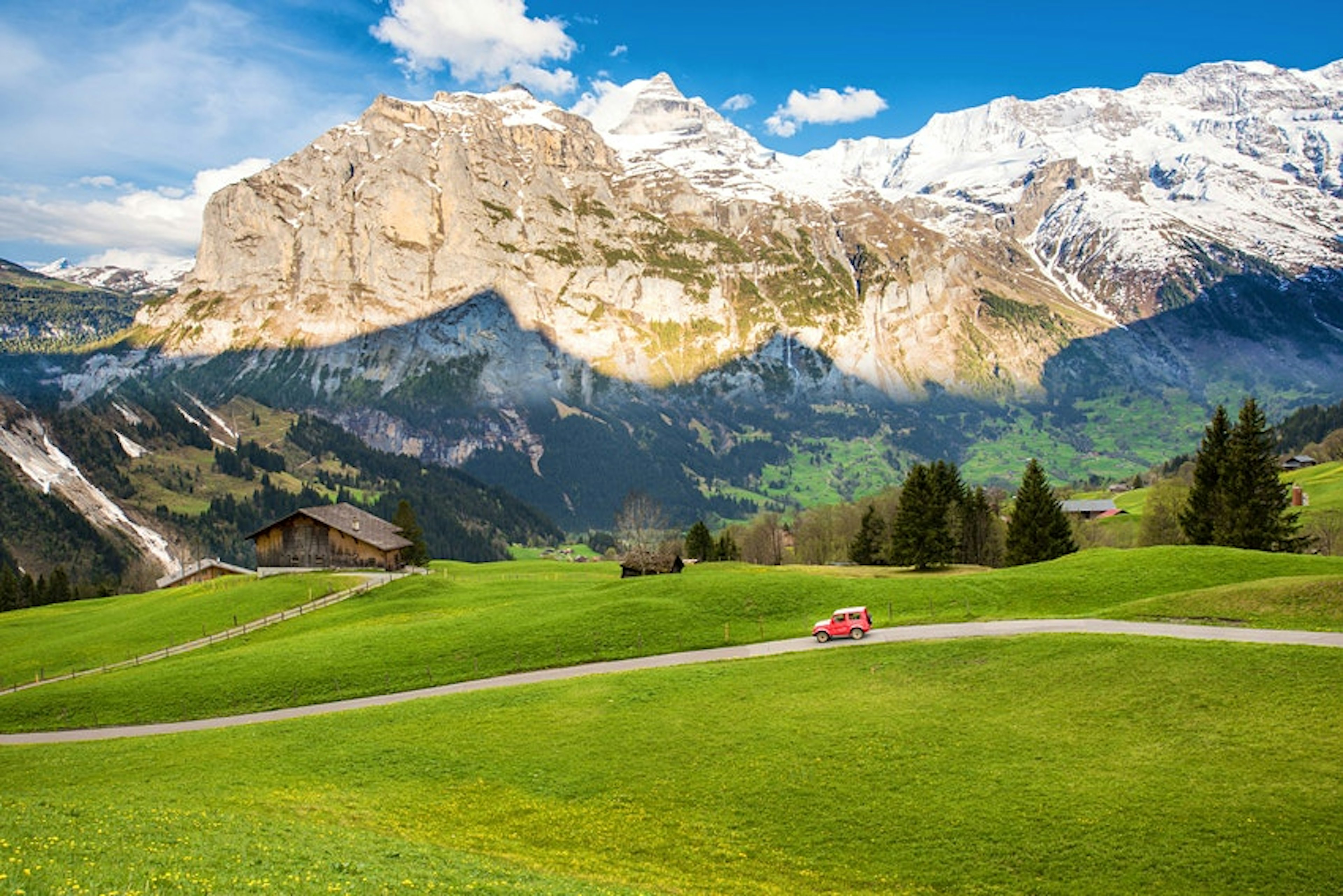 A red car drives down a single lane road along snow-topped mountains in Switzerland