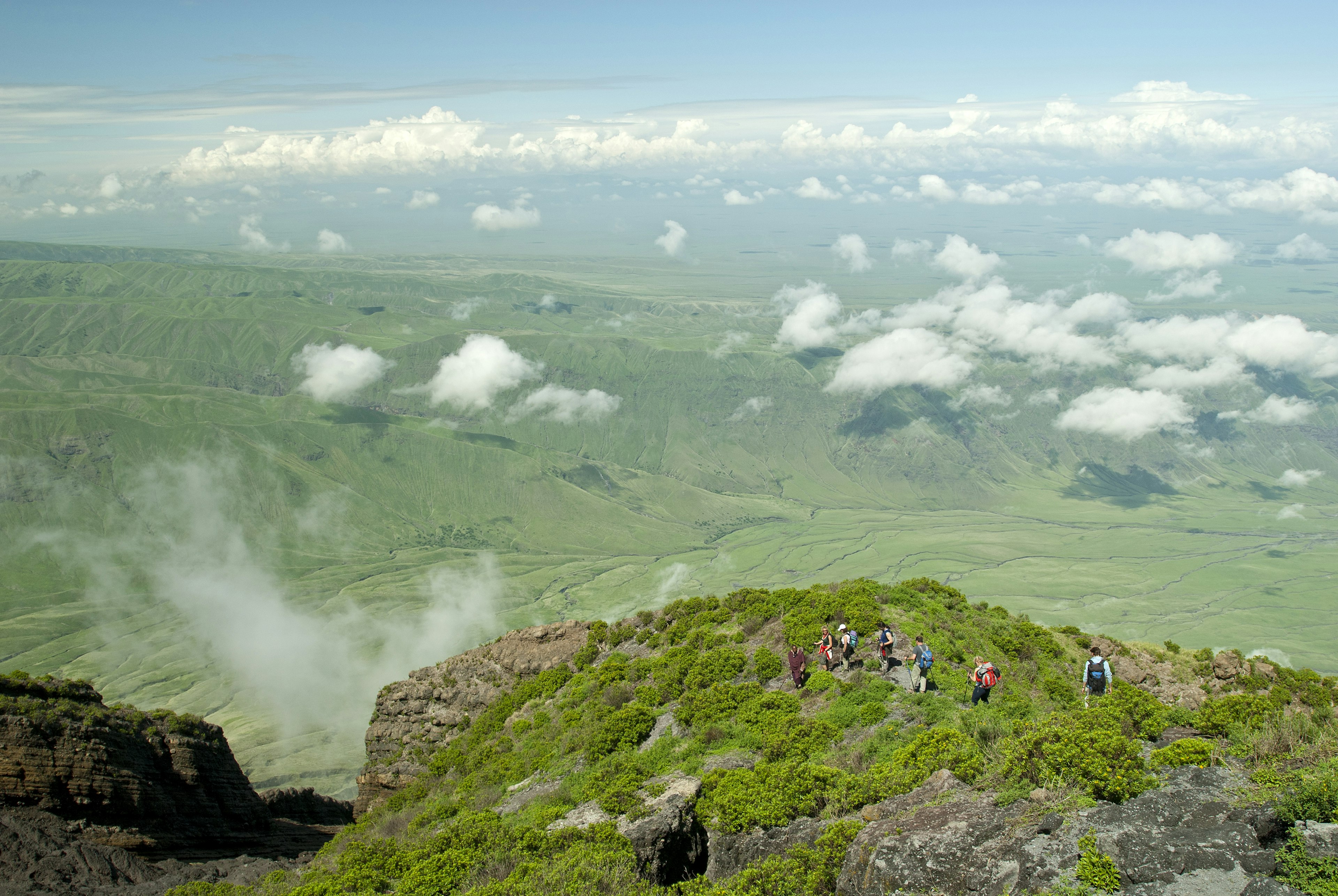 A group hiking down from the top of Ol Doinyo Lengai into the green Rift Valley. In the background the escarpment of the Rift Valley is visible. In the foreground there is some volcanic smoke coming out of a crack.