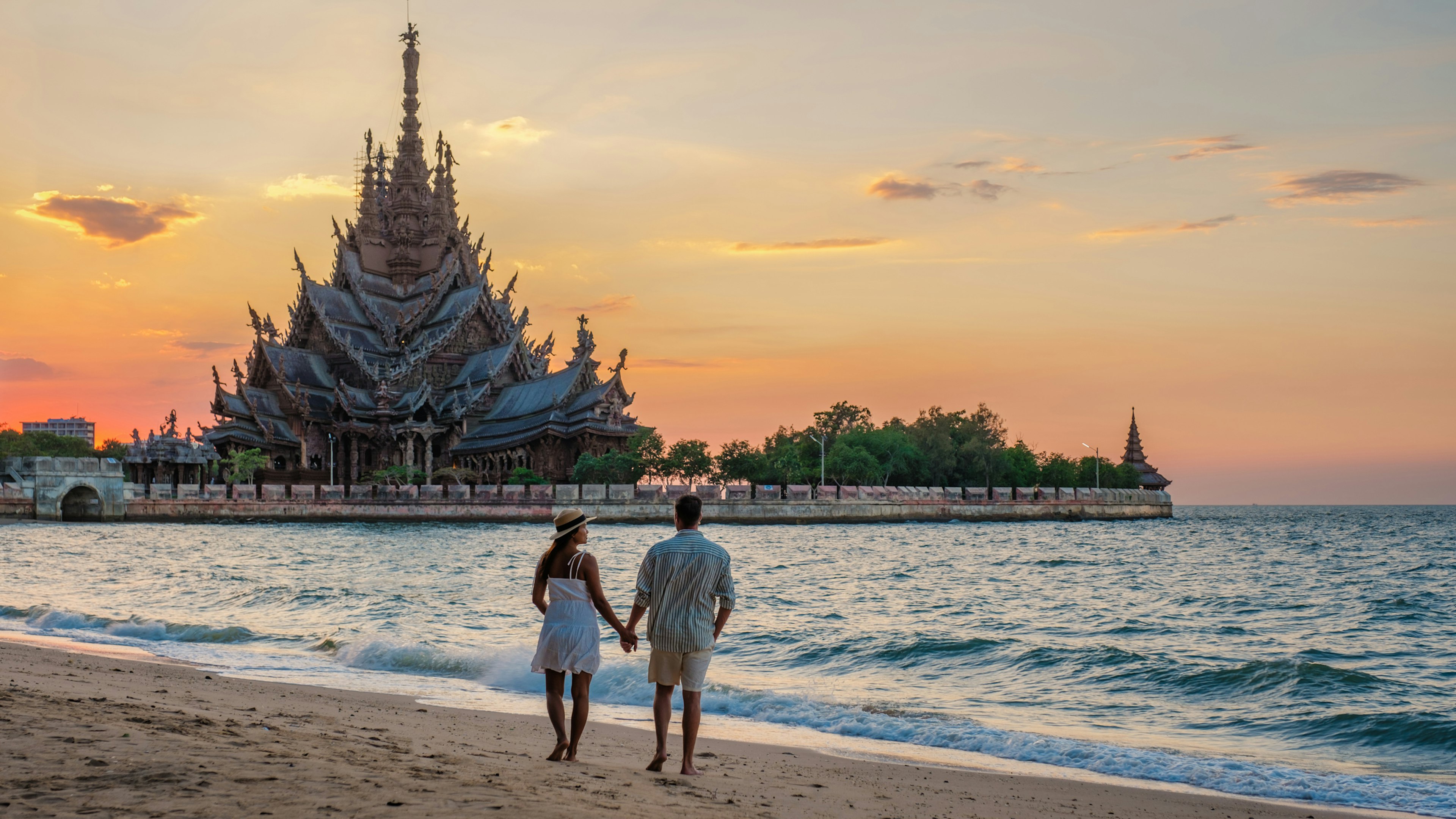 A couple walk on the beach at Pattaya in front of the Sanctuary of Truth temple