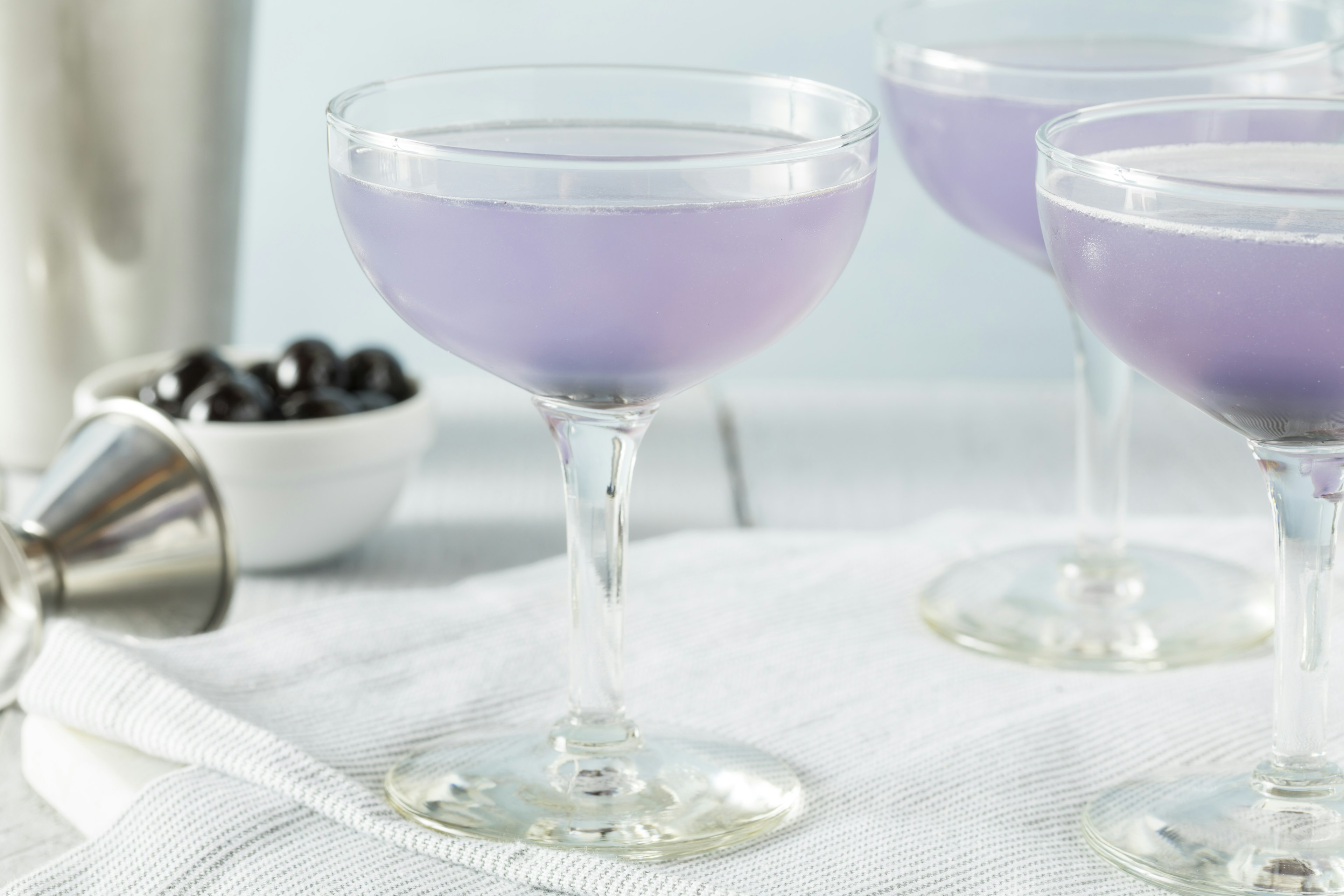 Violet-colored cocktail, the aviation sits on a white cloth. In the background there is a bowl of blueberries.