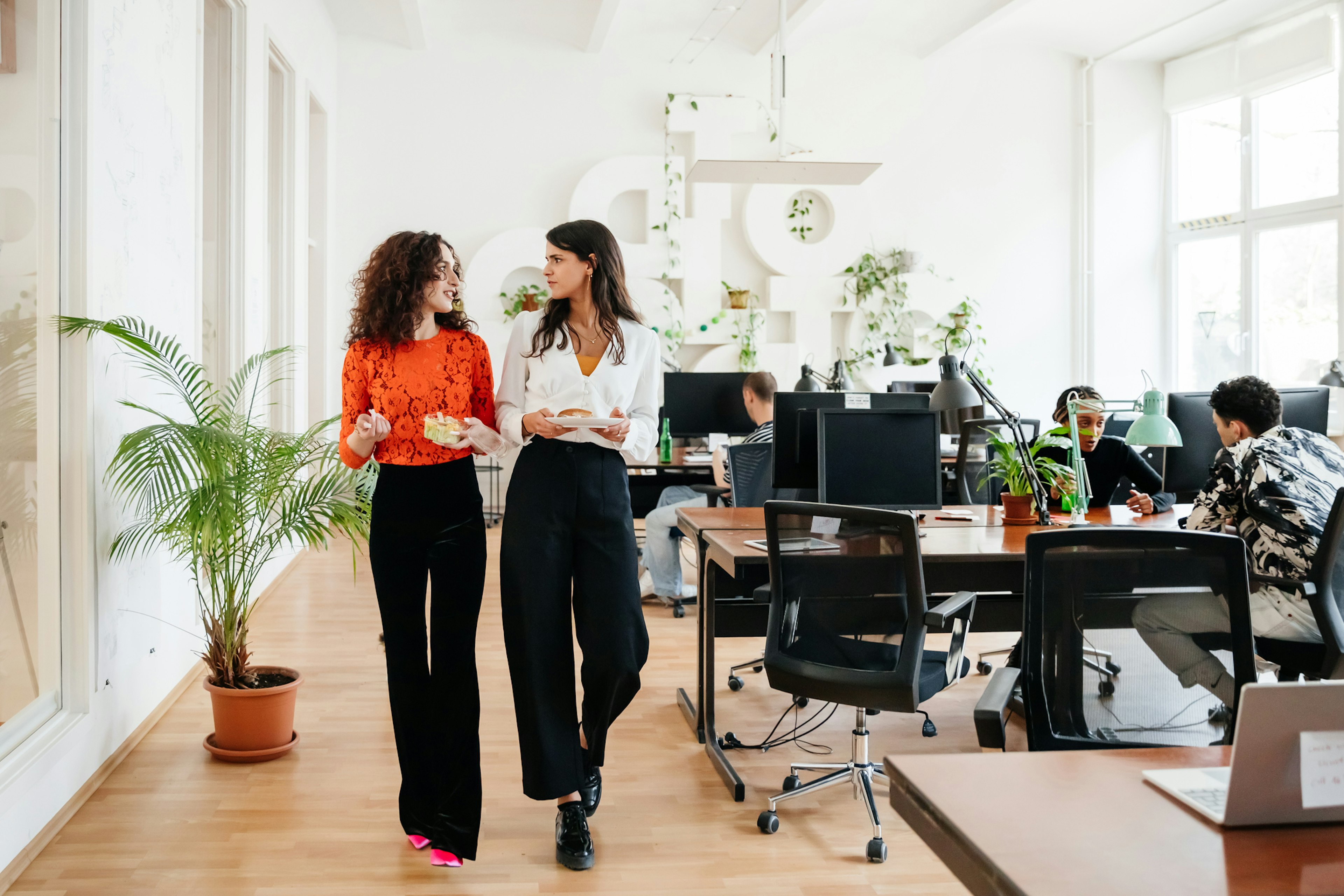 A transgender woman walking past some computer desks heading for her lunch with an office colleague