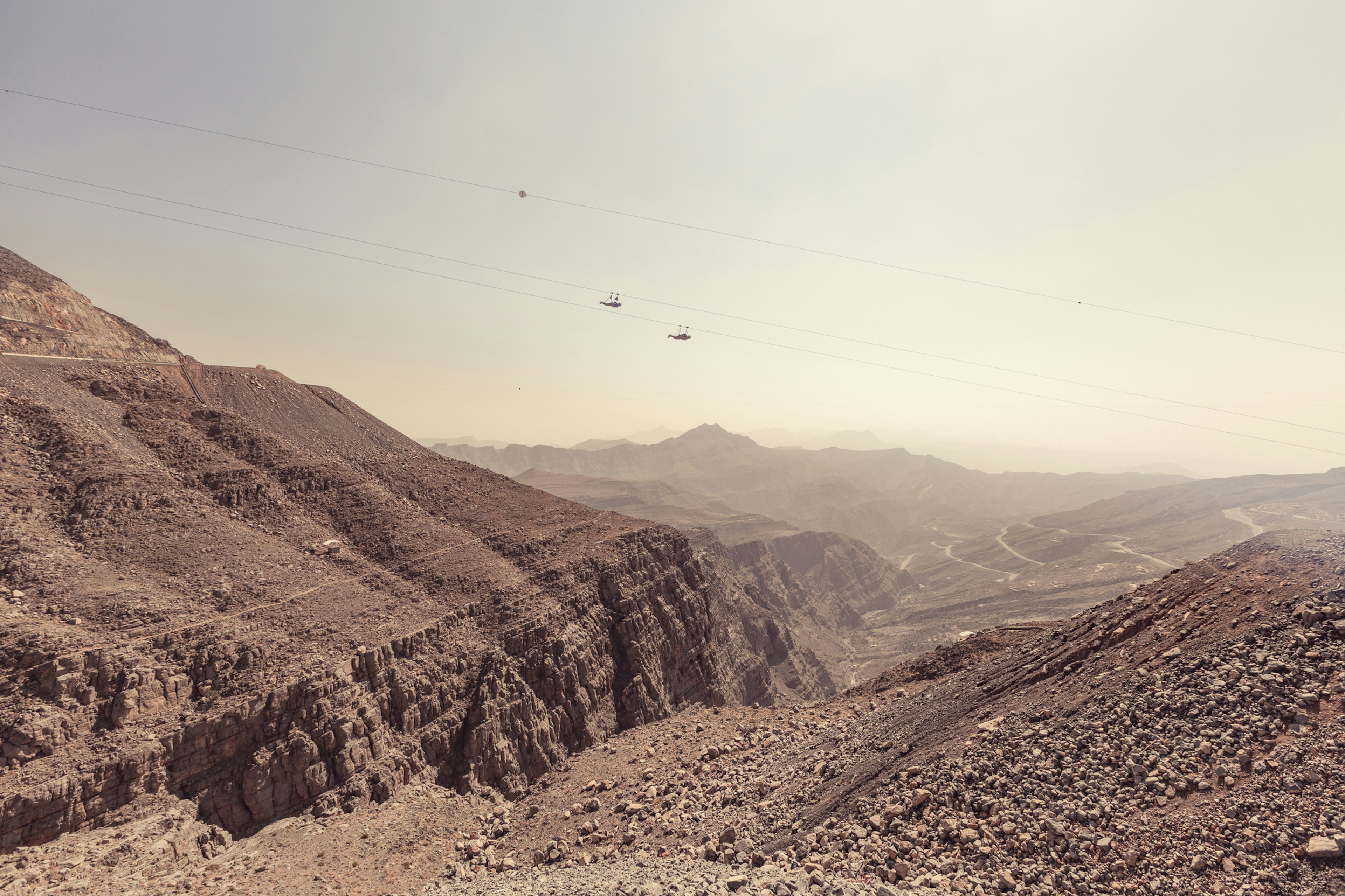 Two zip liners fly over a canyon on wires at a great height above a red rocky landscape