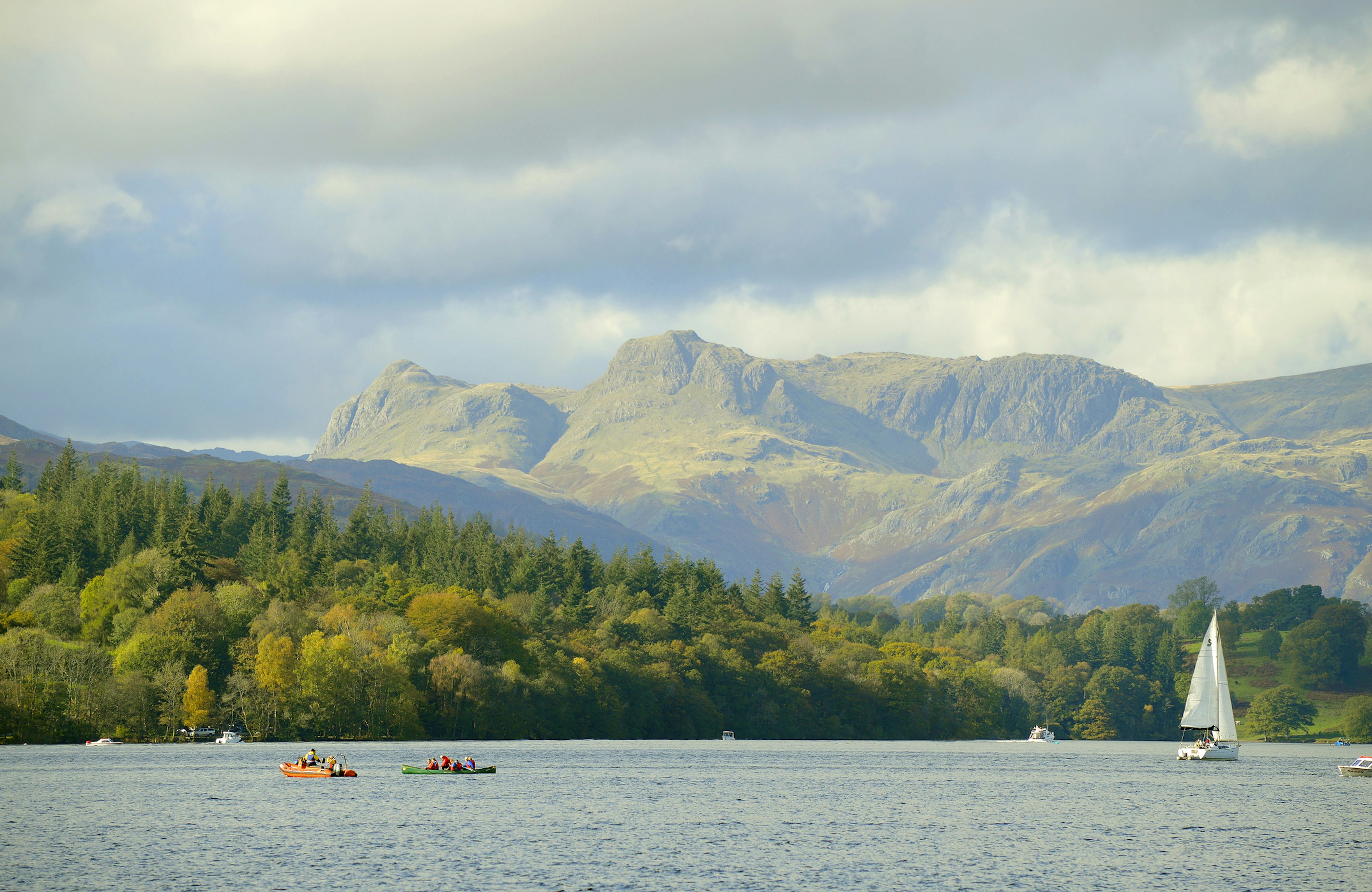 A lake with kayakers out on small boats and a large sail boat with a white sail. It's overlooked by huge green hills in a vibrant national park