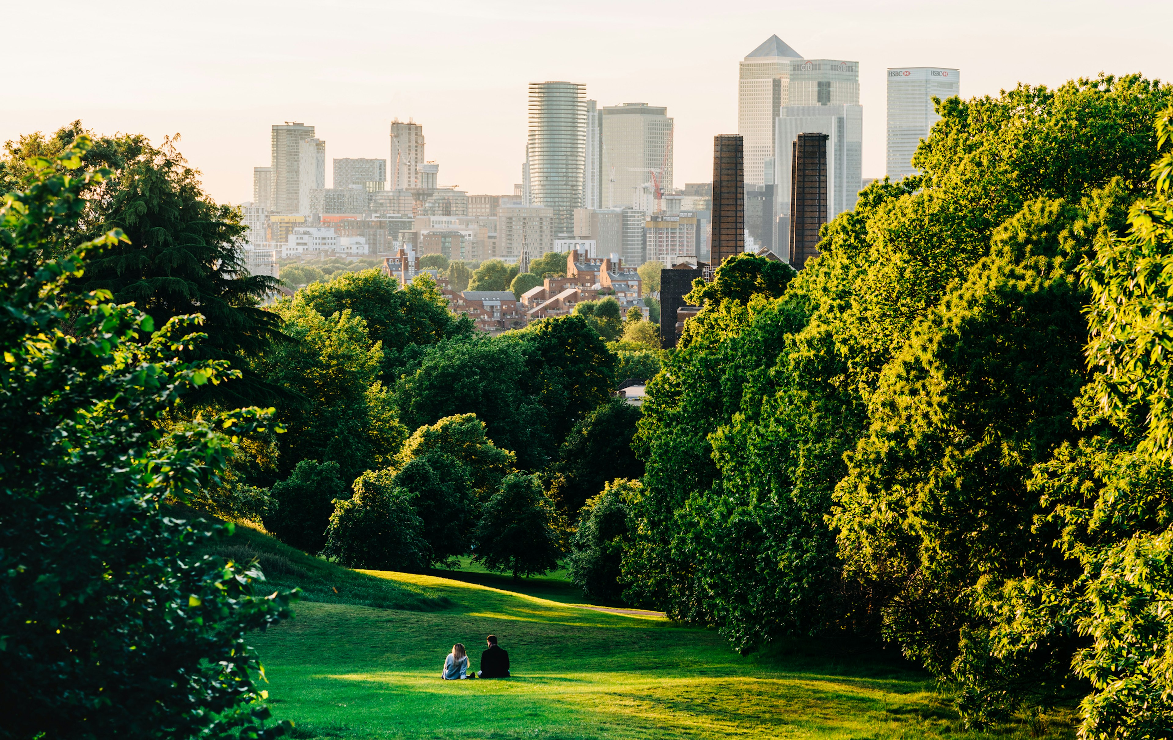 A couple sit in a green field looking towards the Canary Wharf skyline, London.